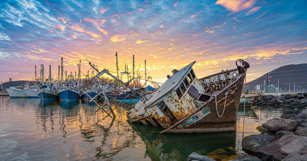 Barco camaronero oxidado y semihundido inclinado en primer plano; al fondo se alinean más embarcaciones pesqueras bajo un cielo de atardecer rosado y dorado reflejado en el agua tranquila del muelle.