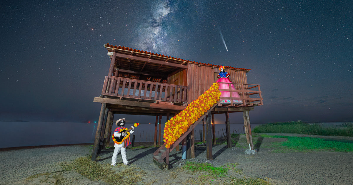 Catrina y catrín frente a un palafito adornado con cempasúchil, bajo la Vía Láctea y un cometa en la playa nocturna.