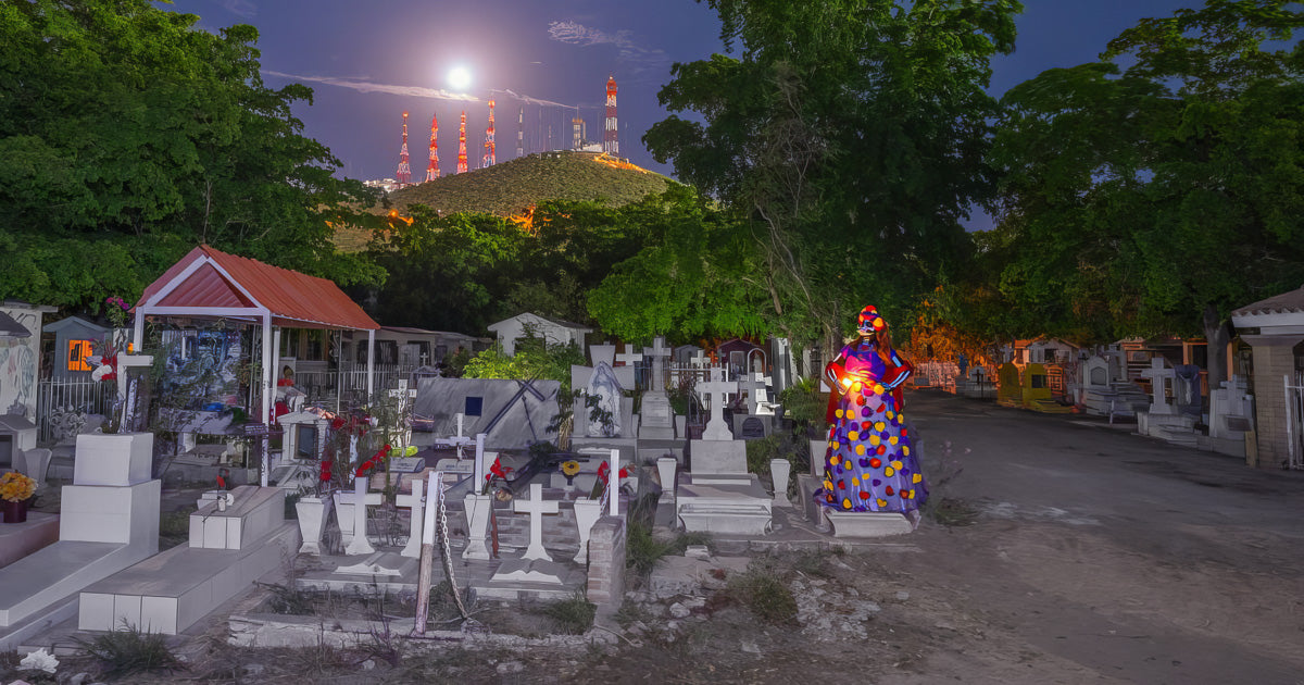 Catrina con vestido colorido sostiene una luz entre cruces y tumbas en un panteón; al fondo se eleva un cerro con antenas bajo la luna llena y un cielo nocturno azul.