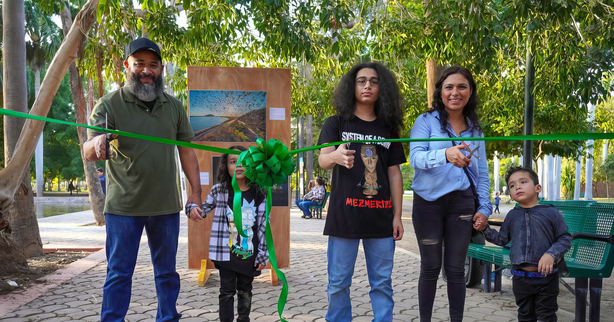 Corte de listón de la exposición fotográfica en el Parque Sinaloa: Edén López y su familia sostienen una cinta verde frente a paneles con imágenes, al aire libre entre árboles.