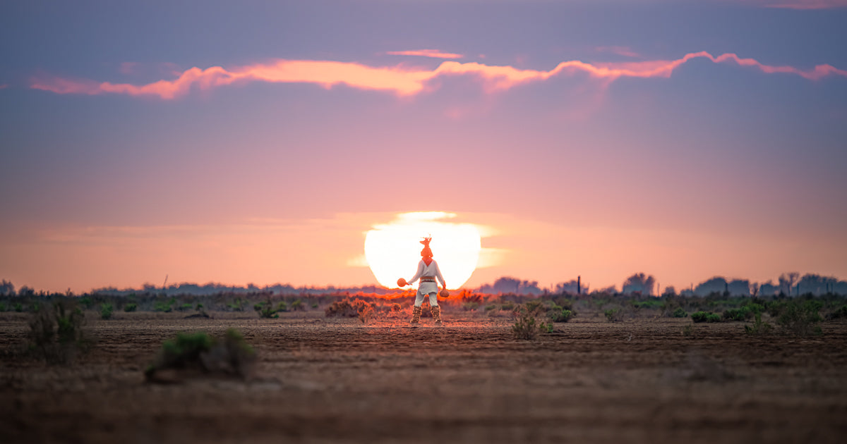 Danzante del venado yoreme centrado frente a un gran sol poniente en planicie semidesértica, cielo rosado y morado.