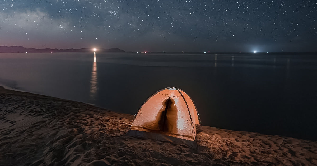 Tienda de campaña iluminada sobre dunas junto al mar en una noche estrellada; silueta dentro de la carpa y reflejos lejanos de luces sobre el agua.