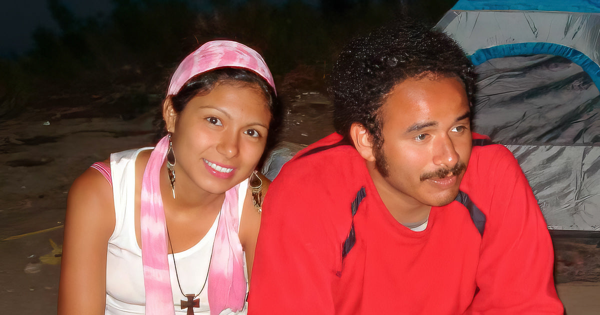 Retrato de dos jóvenes en noche de campamento, sentados junto a una carpa; ella con pañuelo rosa y él con suéter rojo.