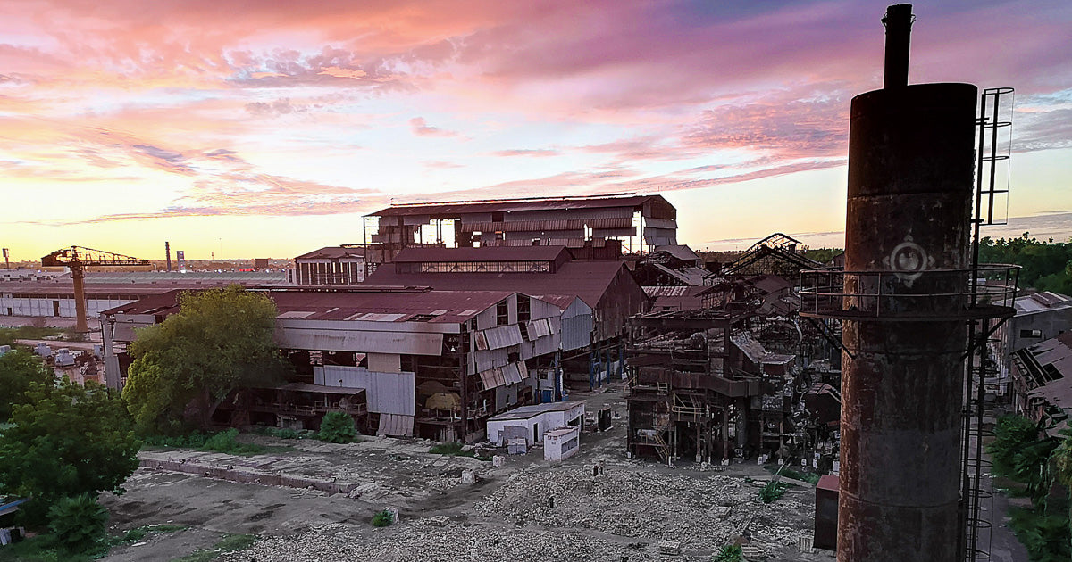 Complejo industrial deteriorado con naves metálicas, gran chimenea a la derecha, cielo rosado de amanecer y vista elevada sobre patios y terreno árido.