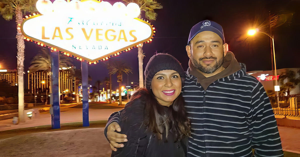 Pareja abrigada posa de noche frente al letrero luminoso de Las Vegas; palmeras y luces de avenida al fondo, encuadre cercano con sonrisa y abrazo.