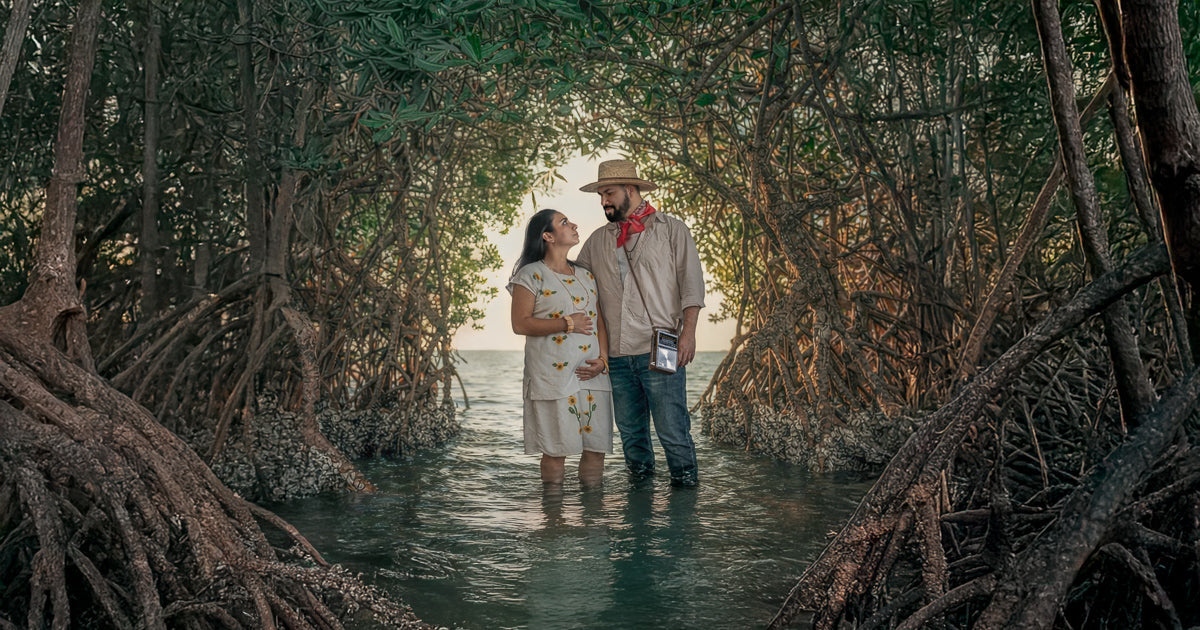 Pareja de pie, con el agua a la altura de las rodillas, dentro de un túnel de manglar; ella embarazada lo mira sonriendo y él la observa con sombrero y paliacate rojo, con el mar al fondo y luz cálida filtrándose entre las ramas.