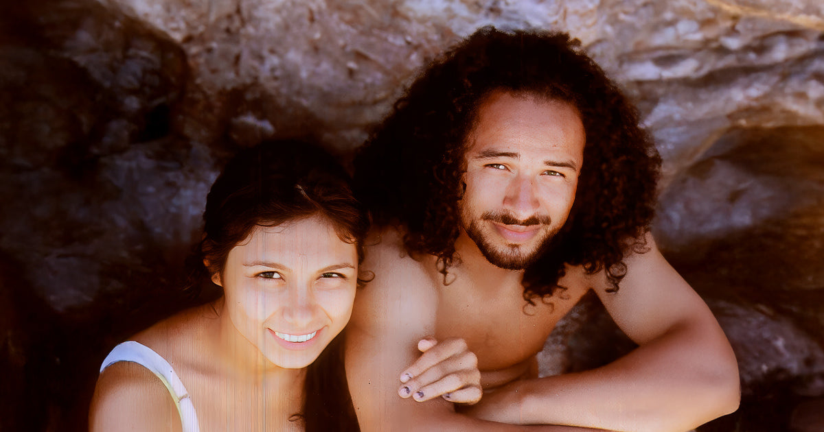 Retrato cercano de una pareja junto a una pared de roca en la costa; luz cálida resalta sus rostros y sonrisa serena, ambiente veraniego al atardecer.