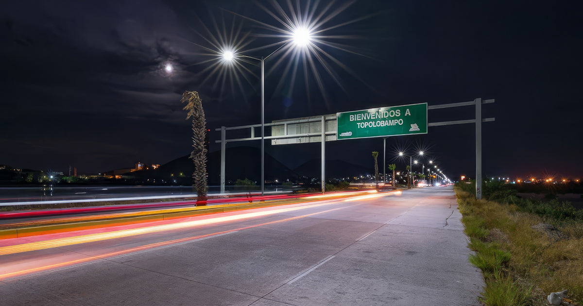 Carretera nocturna con rastro de luces de autos y faroles en estrella junto al letrero “Bienvenidos a Topolobampo”, con la luna entre nubes y cerros al fondo.