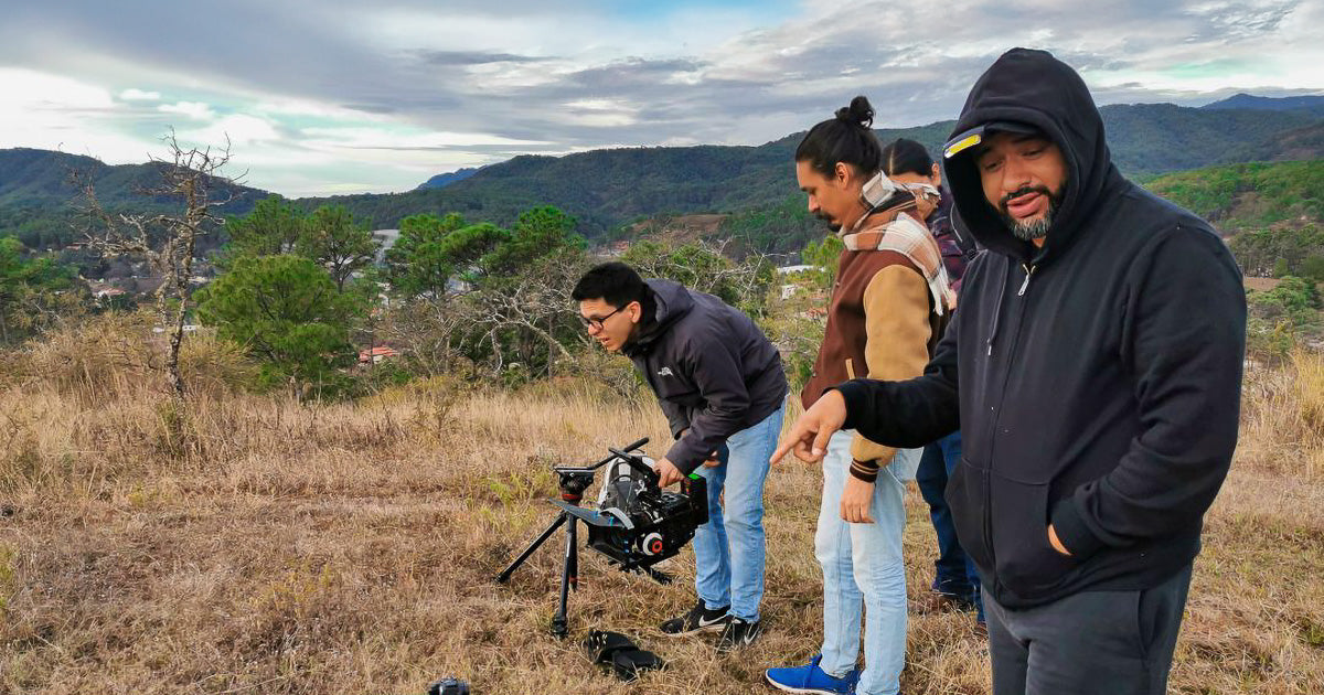 Grupo de personas en una colina con paisaje montañoso; uno señala mientras otro ajusta una cámara de video profesional sobre un soporte en el suelo.