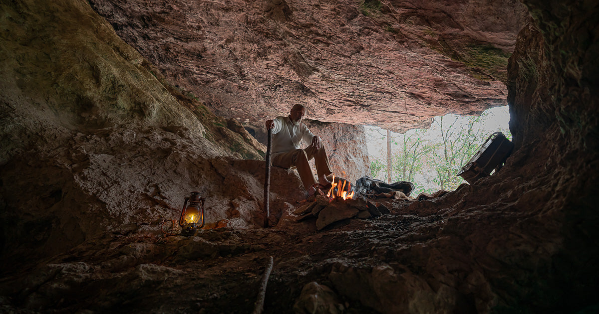 Hombre sentado dentro de una cueva junto a una fogata, con lámpara de aceite y bastón, mientras entra luz diurna por la boca de la cueva con árboles al fondo.