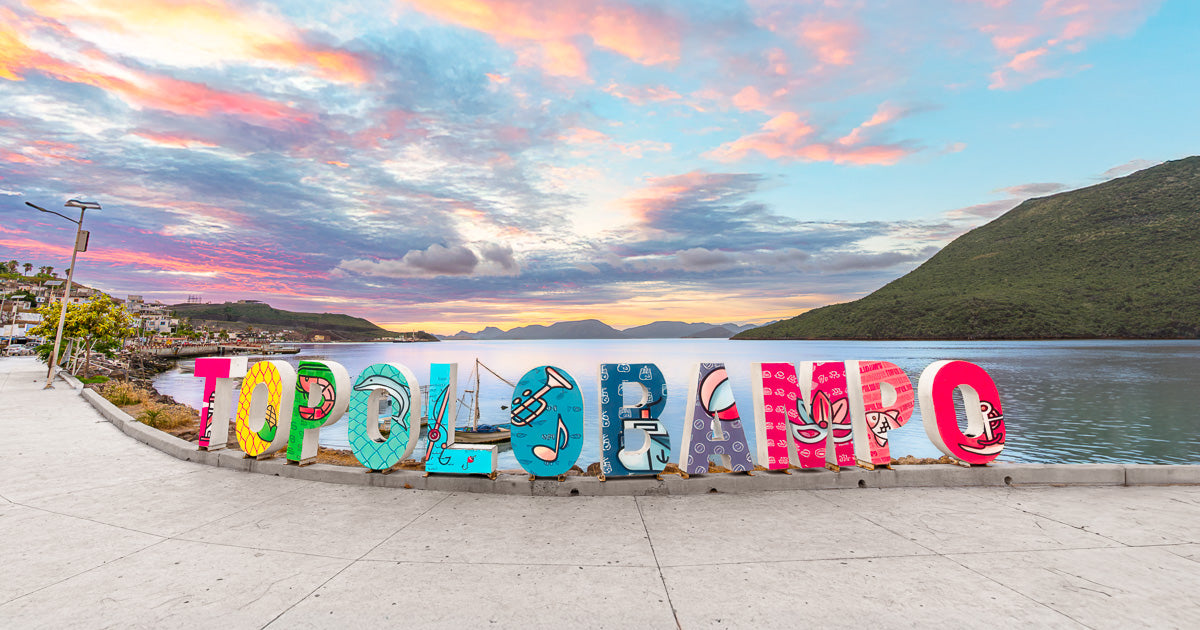 Letras monumentales de “Topolobampo” en el malecón, frente a la bahía y cerros verdes, bajo un cielo de atardecer con nubes rosas y azules.