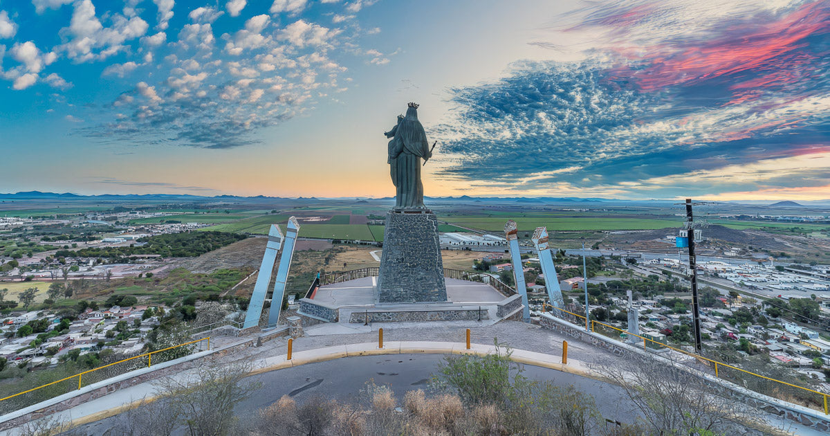 Vista panorámica desde un mirador con una gran estatua religiosa sobre un cerro, campos y ciudad al fondo bajo un cielo con nubes coloridas al atardecer.