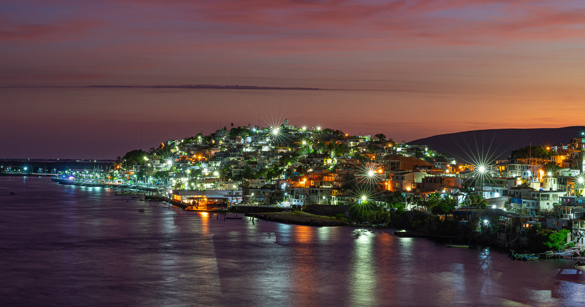 Vista nocturna de un poblado costero sobre una colina, con luces en forma de estrella y reflejos en el agua bajo un cielo magenta y naranja al atardecer.