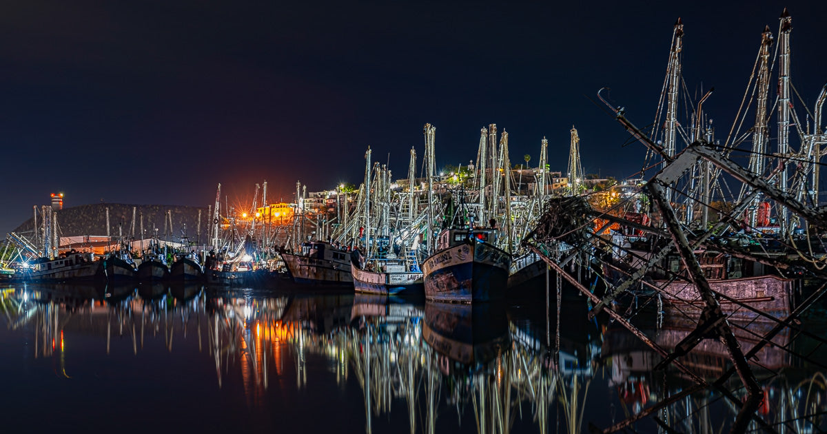 Flota pesquera iluminada atracada de noche; numerosos mástiles y aparejos se reflejan en el agua tranquila del muelle bajo un cielo oscuro.