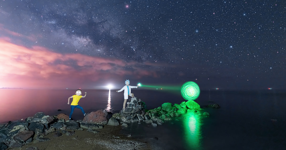 Noche estrellada con la Vía Láctea sobre el mar; Rick y Morty aparecen iluminados sobre rocas junto a un “portal” verde de luz reflejado en el agua.
