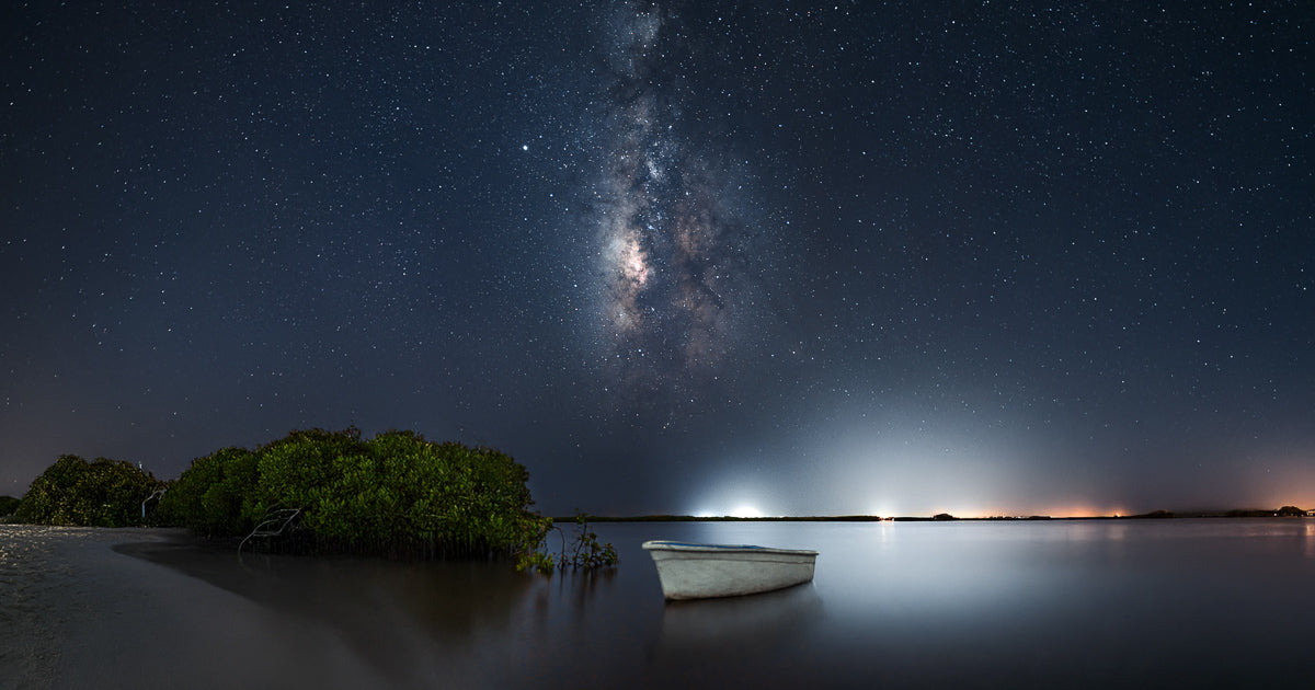 La Vía Láctea vertical sobre un manglar y una pequeña lancha en aguas tranquilas; cielo oscuro lleno de estrellas y resplandor lejano en el horizonte, larga exposición en la laguna.