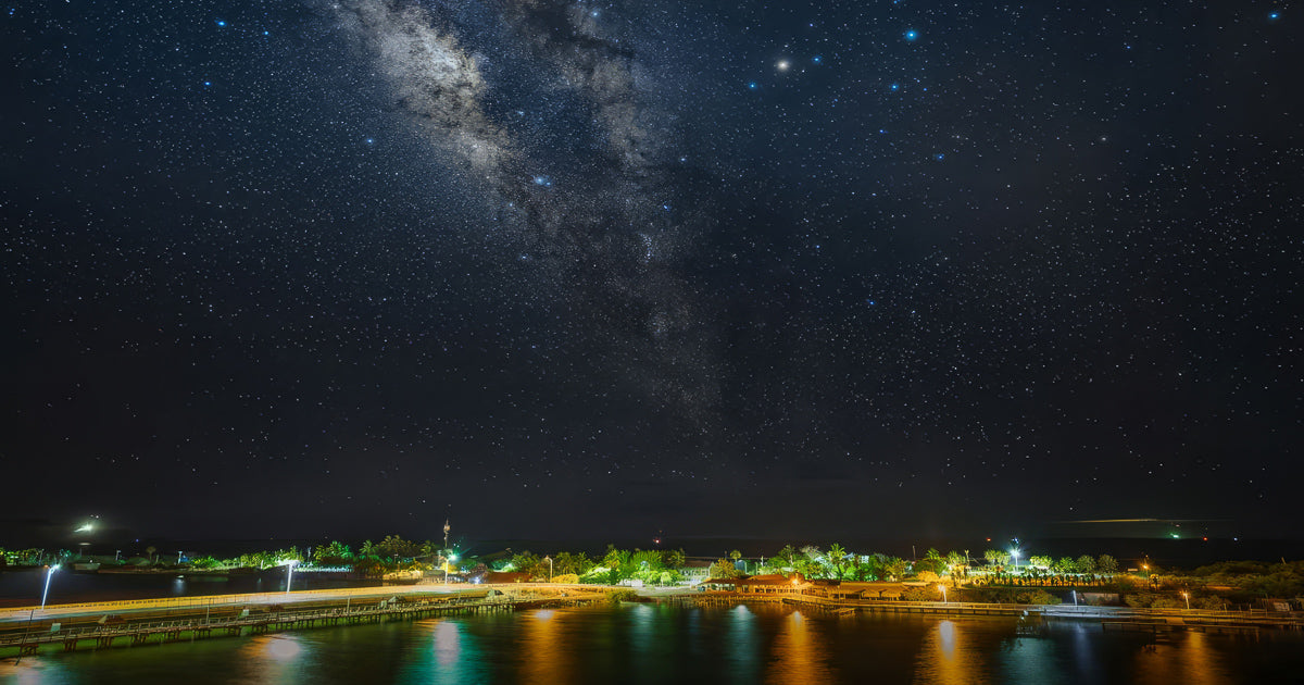 La Vía Láctea sobre un cielo estrellado en Topolobampo, con el malecón y un muelle iluminado reflejándose en el agua.