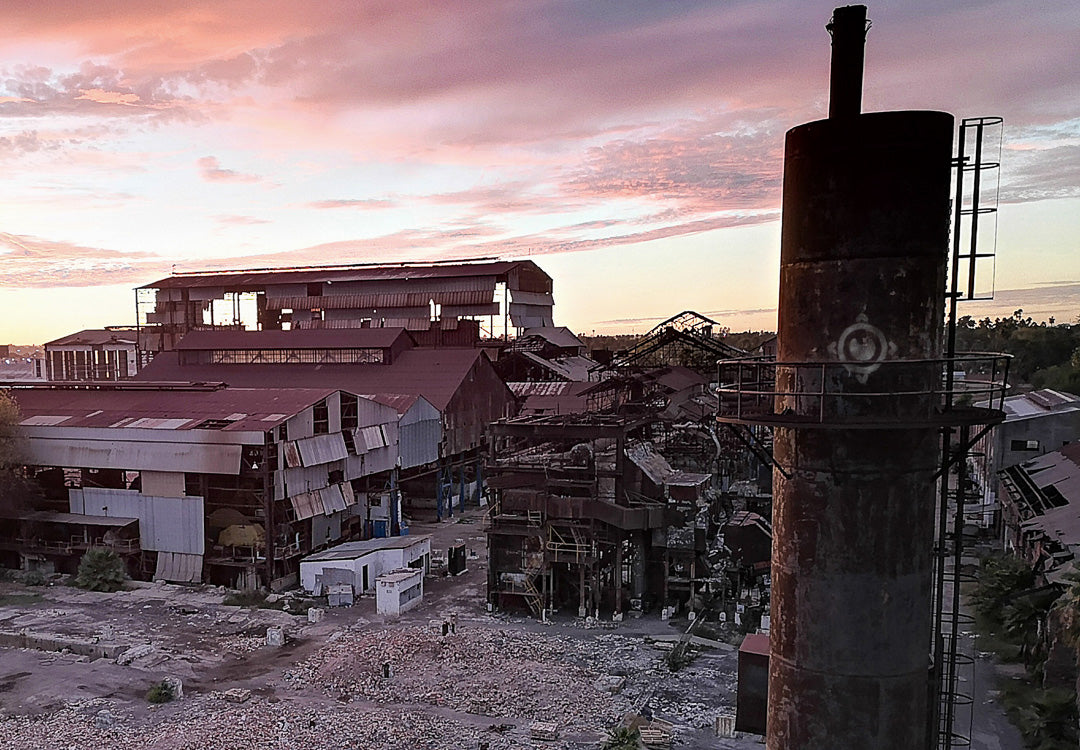 Conjunto de naves industriales deterioradas y chimenea cilíndrica en primer plano; cielo rosado al amanecer sobre el sitio.