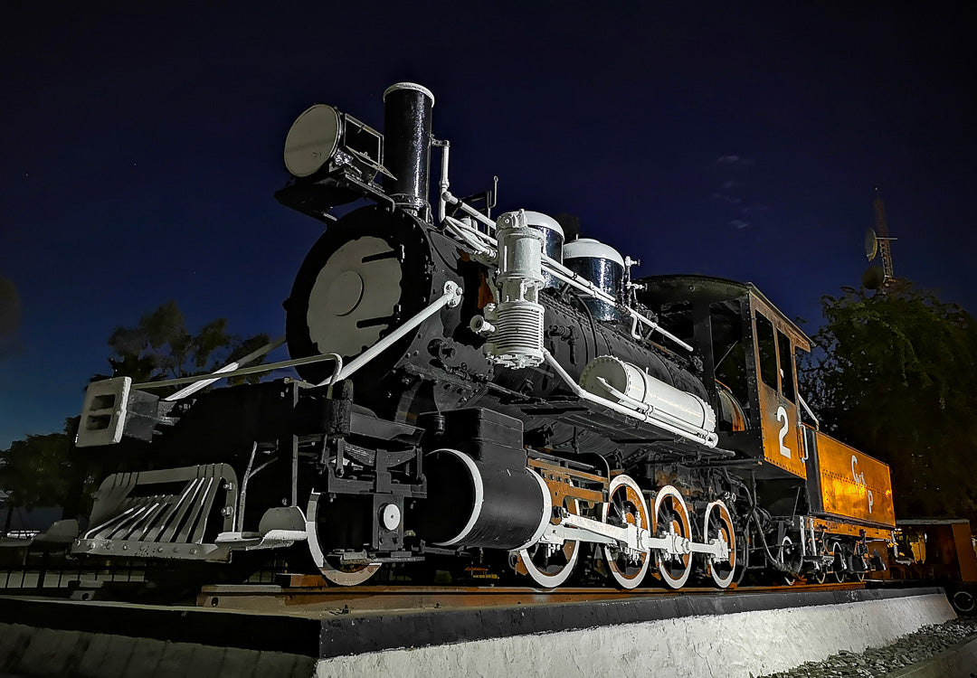 Locomotora de vapor preservada sobre base de concreto; vista en contrapicado durante la noche.