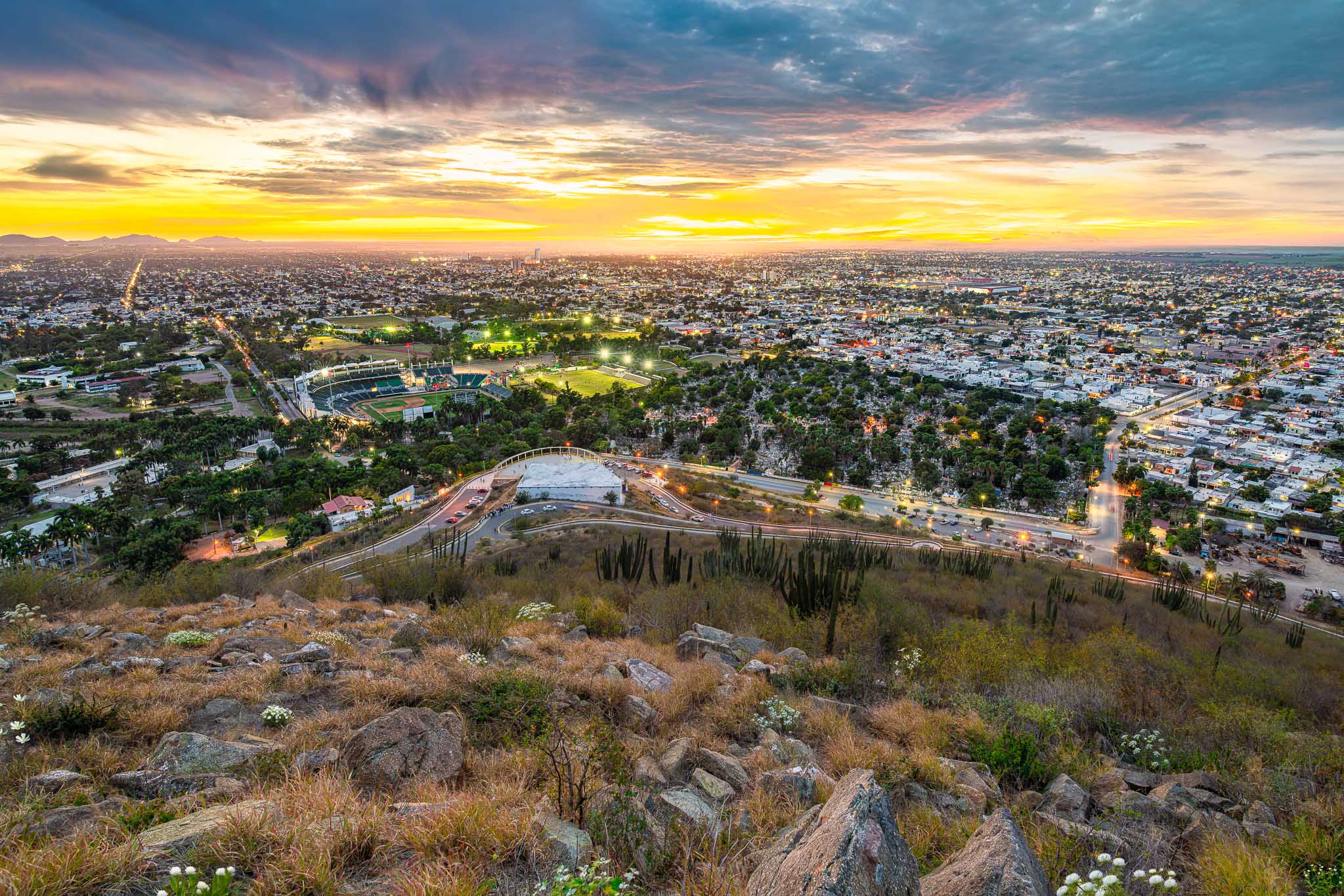 Vista panorámica desde ladera rocosa; ciudad encendida al atardecer, estadio iluminado y avenidas, cielo con nubes doradas.