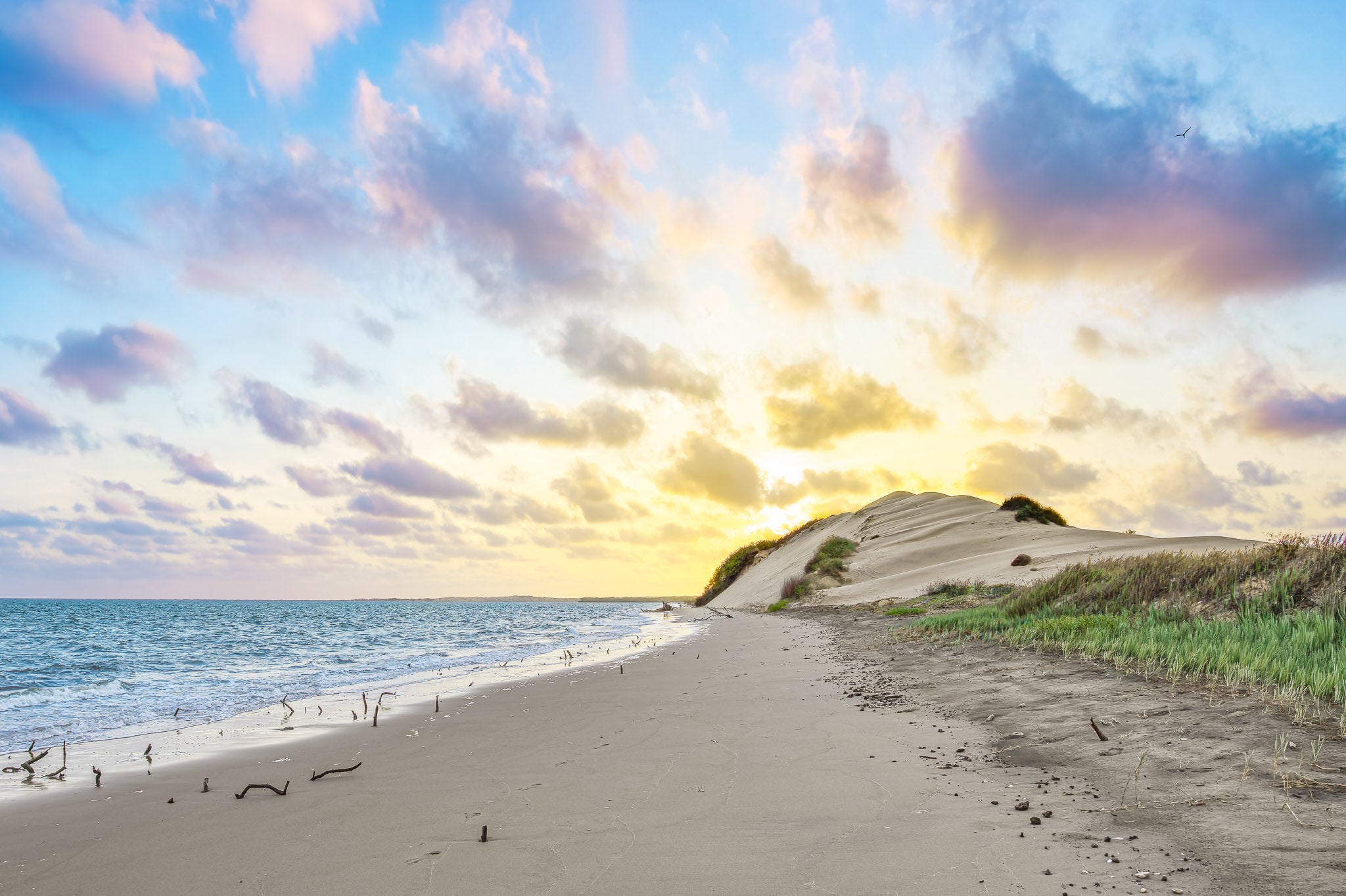Paisaje de playa con gran duna iluminada por el sol naciente; océano a la izquierda, arena con huellas y nubes rosadas abiertas en el horizonte.