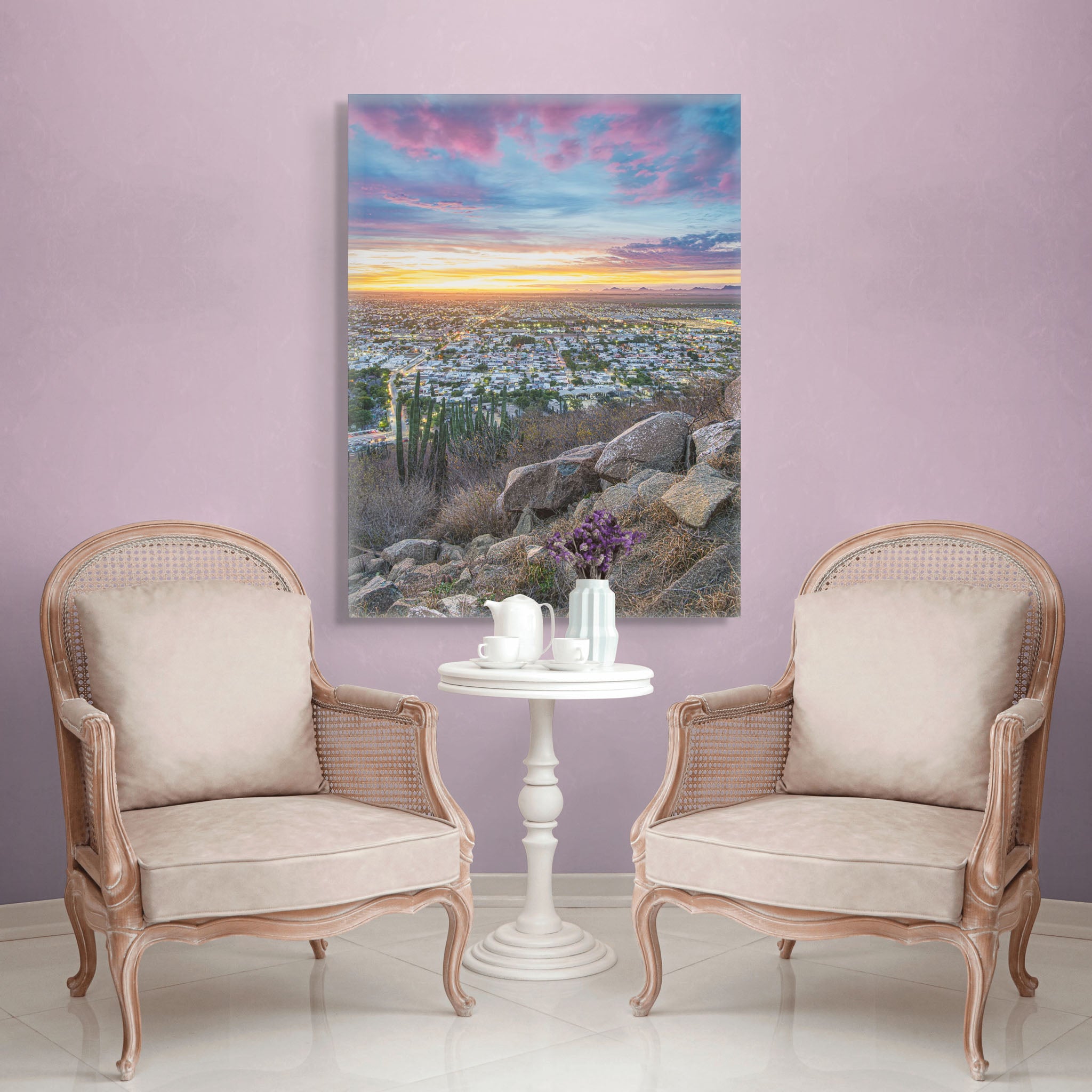 Canvas en muro neutro; vista urbana al atardecer desde altura, cielo magenta y dorado, rocas y cactus en primer plano; atmósfera serena y luminosa.