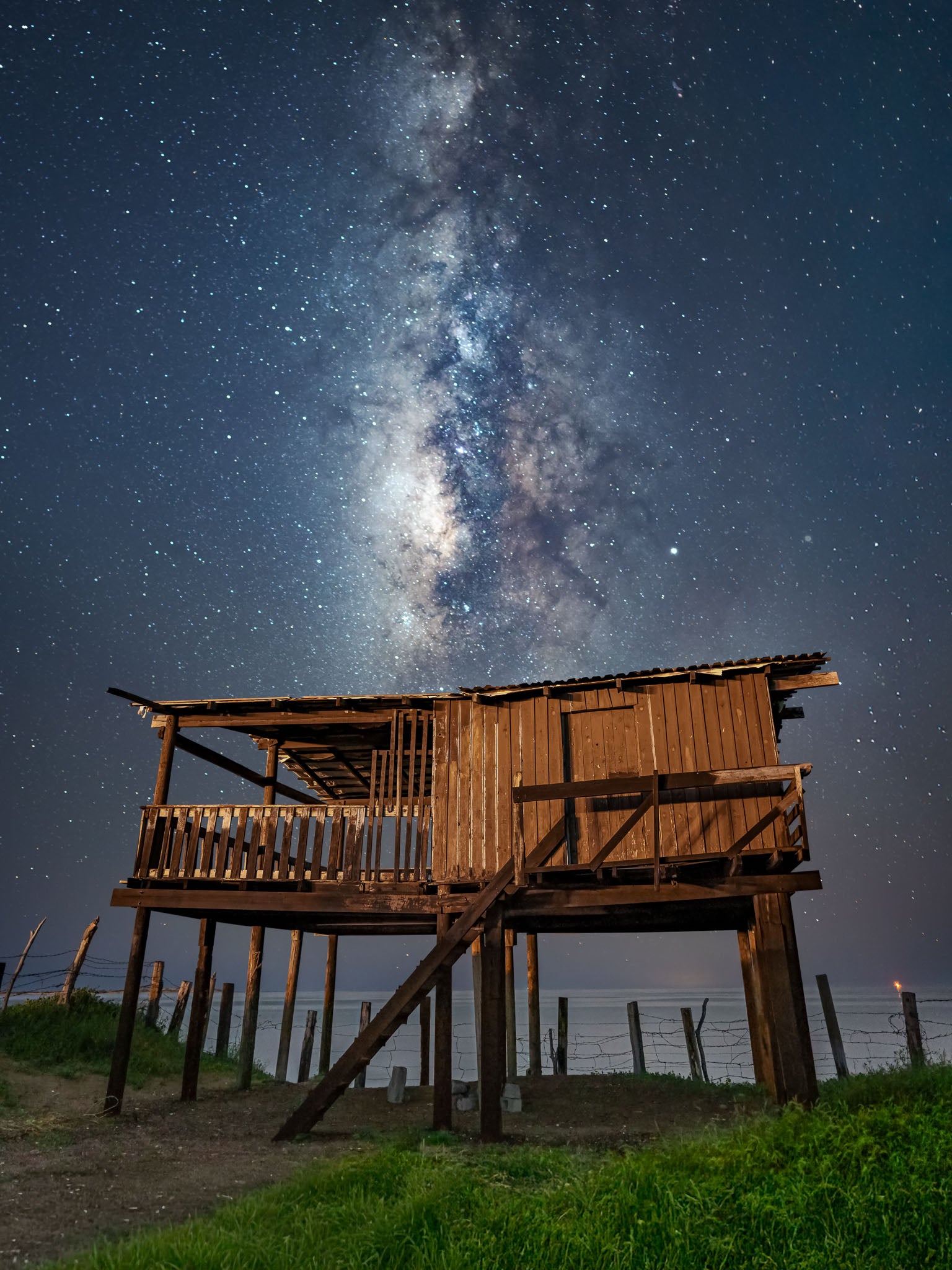 Palafito de madera iluminado frente al mar; vía láctea vertical al fondo, cielo densamente estrellado, nocturna.