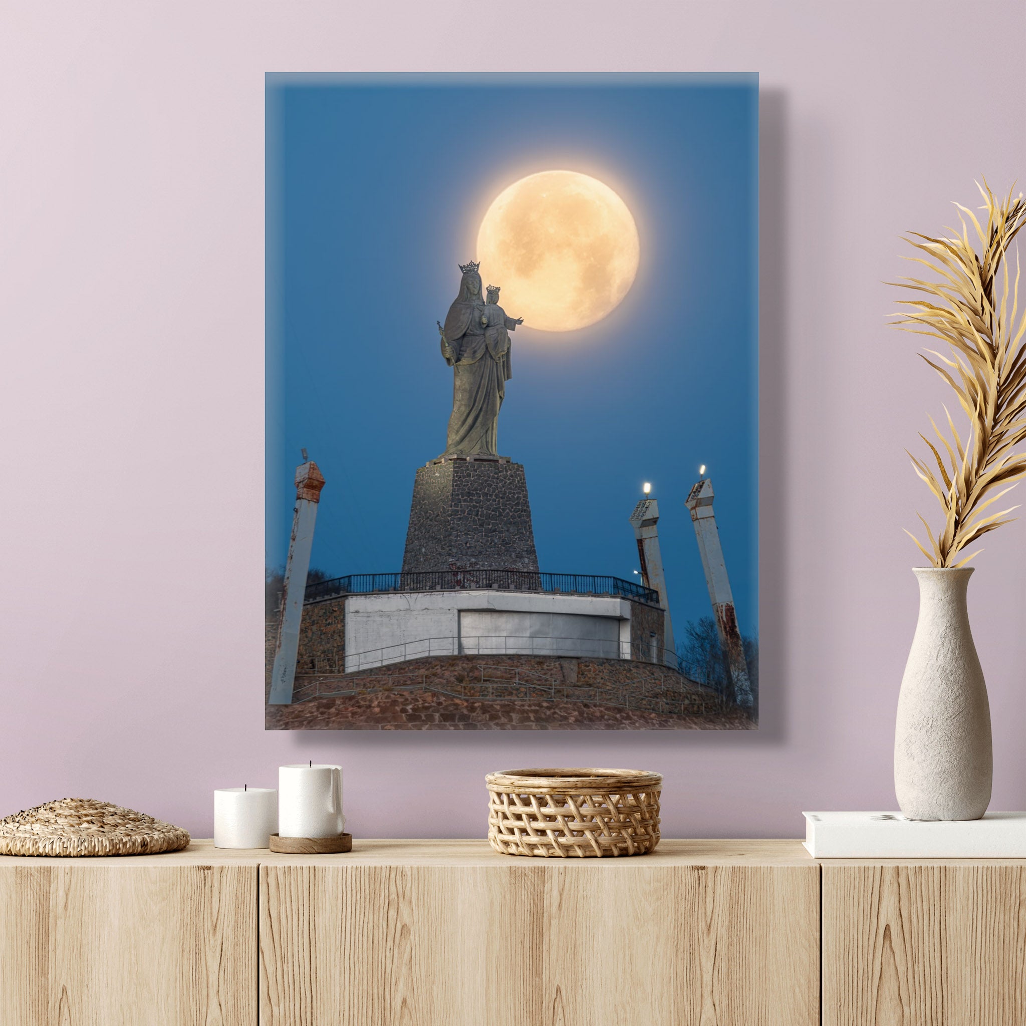 Lienzo vertical en un interior sobrio. La obra muestra la luna llena posada sobre el hombro de la escultura de la Virgen en un cerro.