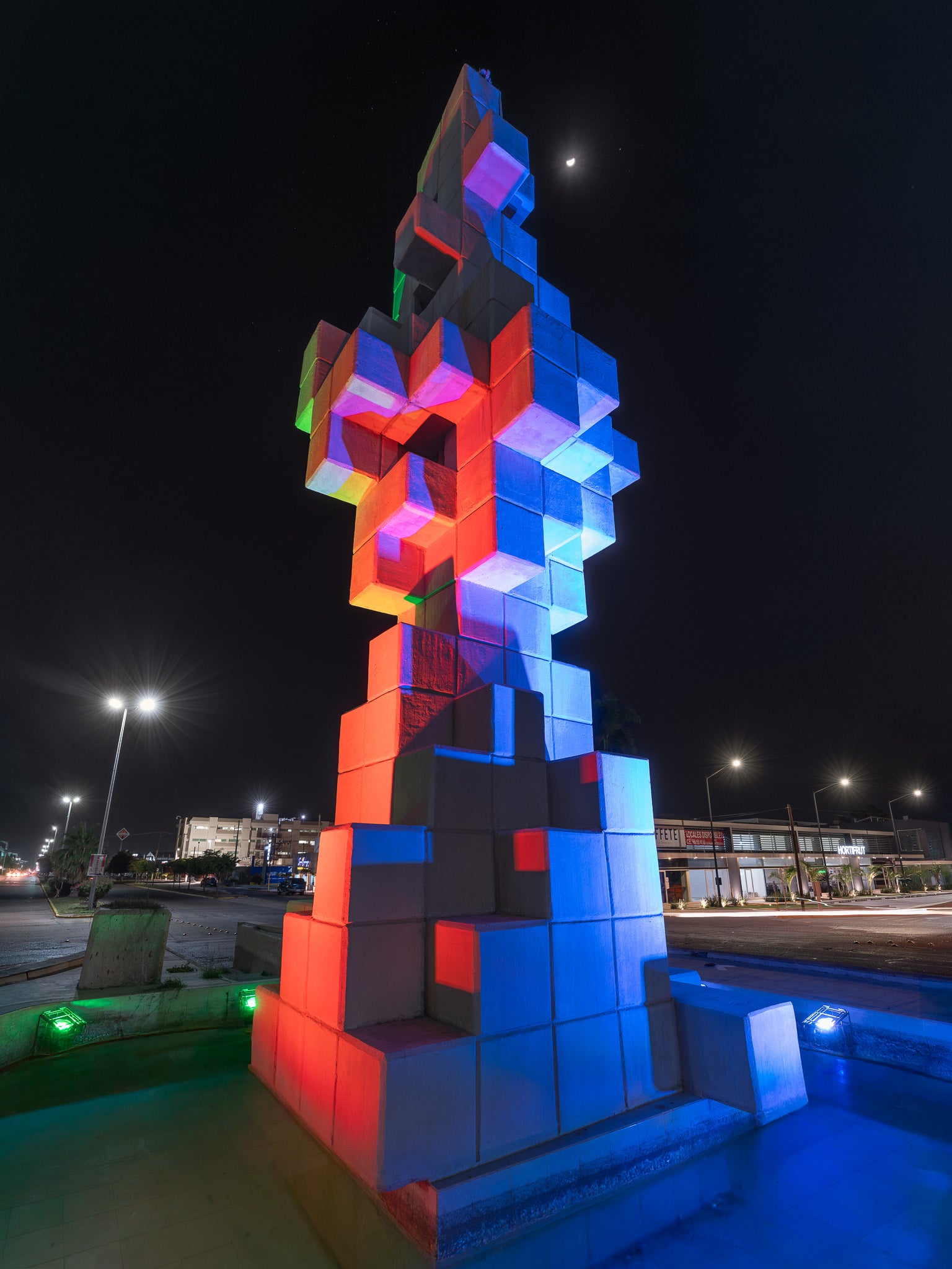 Escultura de cubos de concreto iluminada en azul y rojo, vista en contrapicado nocturno; farolas y luna creciente resaltan el cielo oscuro.