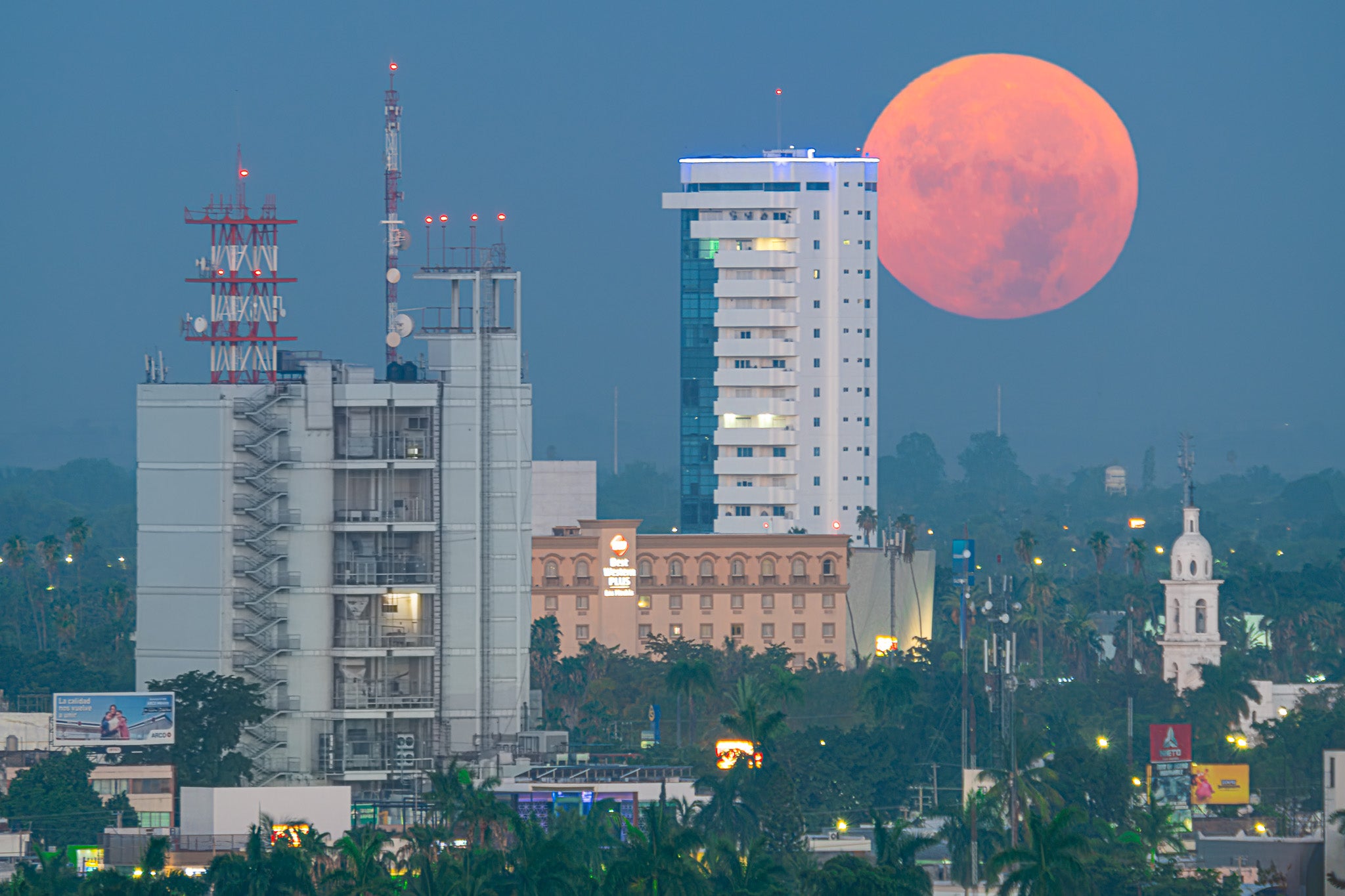 Luna llena anaranjada asoma tras torres y antenas urbanas; palmeras y luces tenues en hora azul, panorámica horizontal.