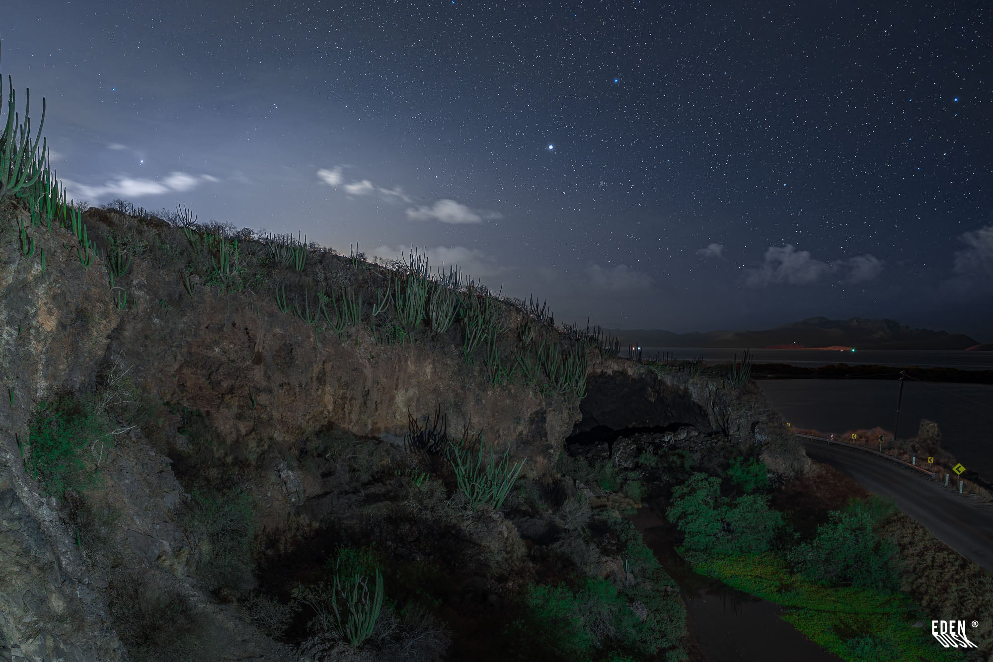 Acantilado con entrada de cueva y cactus bajo cielo estrellado, con camino y laguna al fondo, Cueva de los Murciélagos, El Maviri, Sinaloa.