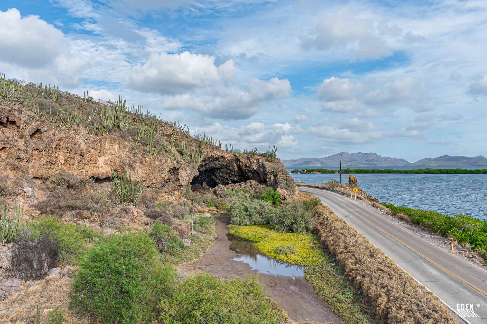 Boca de la Cueva de los Murciélagos junto a carretera y laguna con manglar, cactus en la ladera y cielo con nubes, El Maviri, Sinaloa.