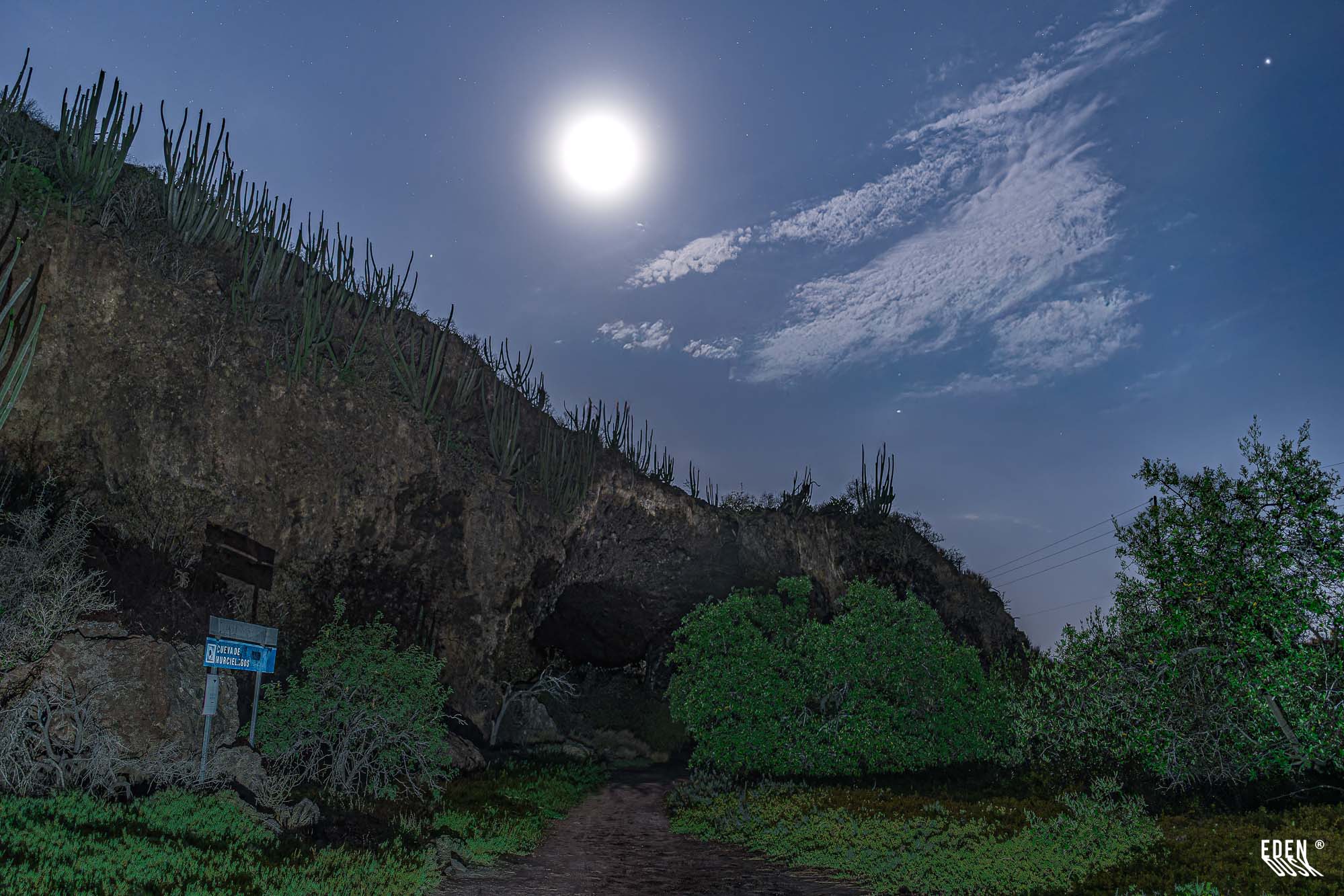 Entrada de la Cueva de los Murciélagos bajo luna brillante, cactus en el cerro, nubes y manglar en primer plano, El Maviri, Sinaloa.