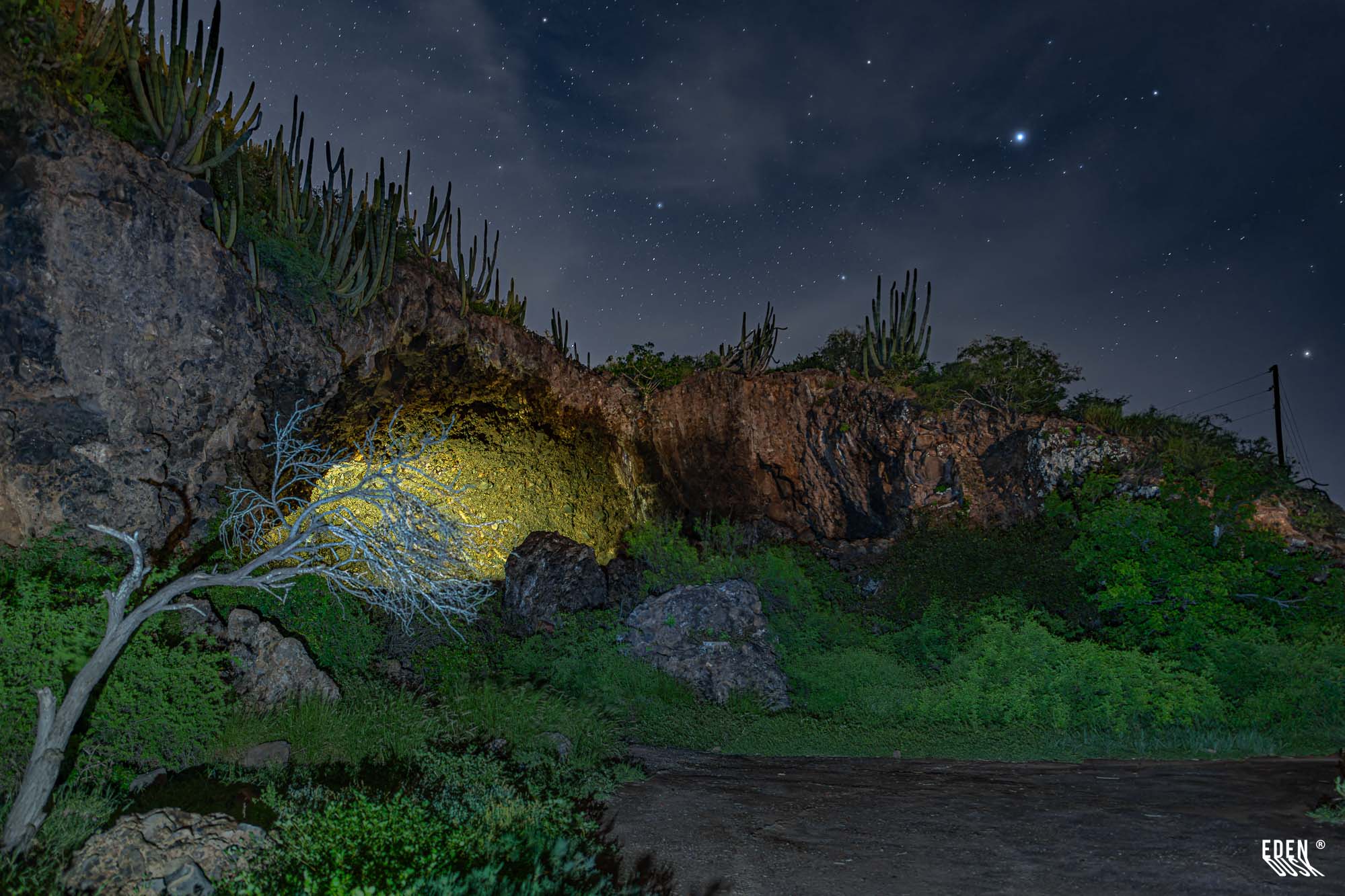 Entrada de la Cueva de los Murciélagos iluminada, cerro con cactus y cielo estrellado, El Maviri, Sinaloa.