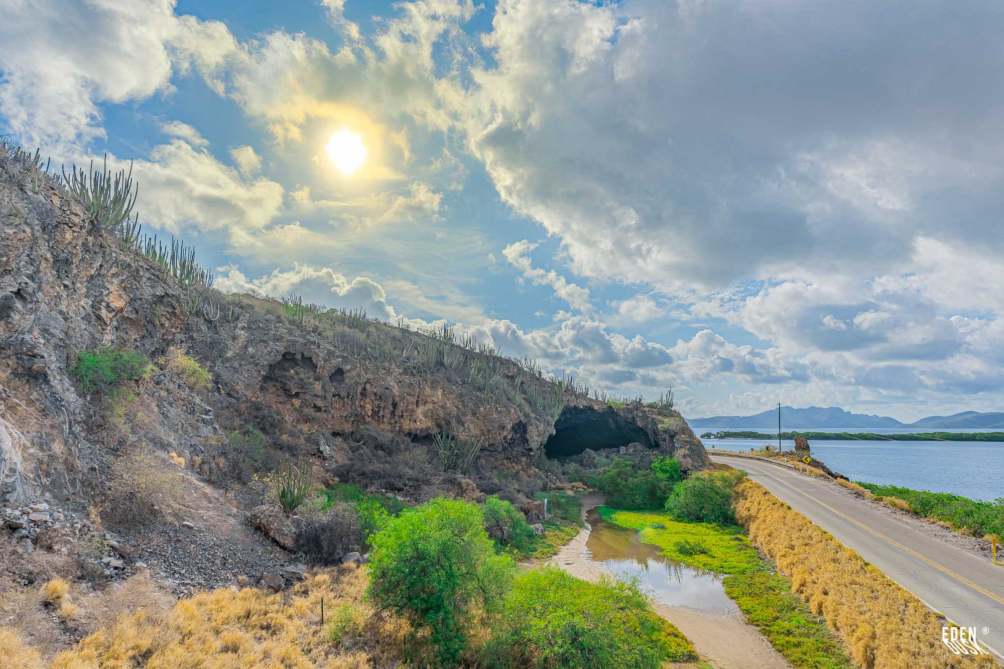 Entrada de la Cueva de los Murciélagos a plena luz del día, cielo con nubes dramáticas, carretera costera y canal de marea junto al manglar y la laguna, El Maviri, Sinaloa.