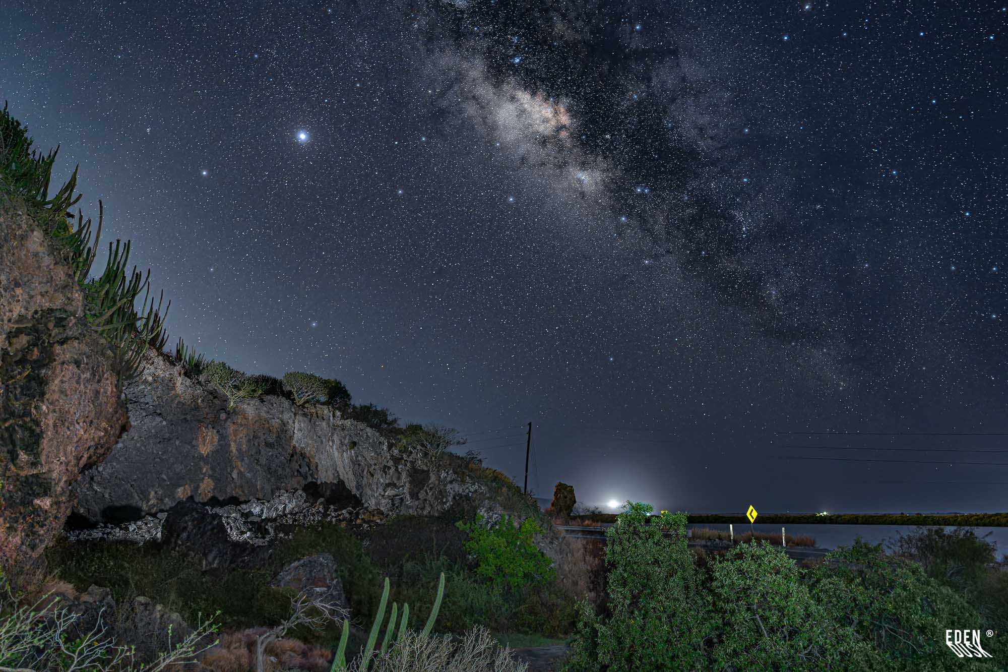 Vía Láctea nítida sobre la entrada rocosa de la Cueva de los Murciélagos, con cactáceas, curva de carretera y resplandor lejano junto a la laguna, El Maviri, Sinaloa.