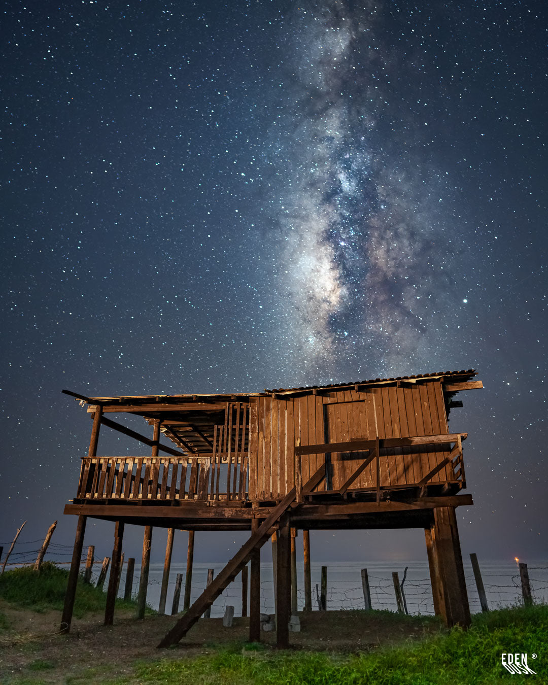 Cabaña de madera elevada sobre pilotes frente al mar, con la Vía Láctea ascendiendo luminosa en el cielo nocturno; cerca de alambre al fondo y luz cálida iluminando la estructura en El Maviri.