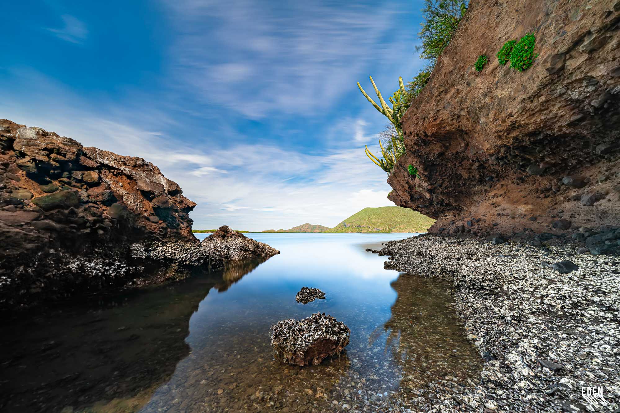 Cala estrecha entre rocas oscuras y agua tranquila; pared con cactus a la derecha y colinas bajas al fondo bajo cielo claro.