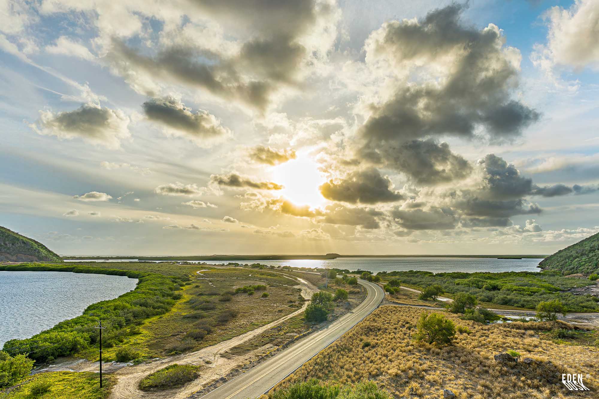 Carretera curva entre matorrales y lagunas; sol bajo entre nubes con rayos al horizonte y cerros a los lados.