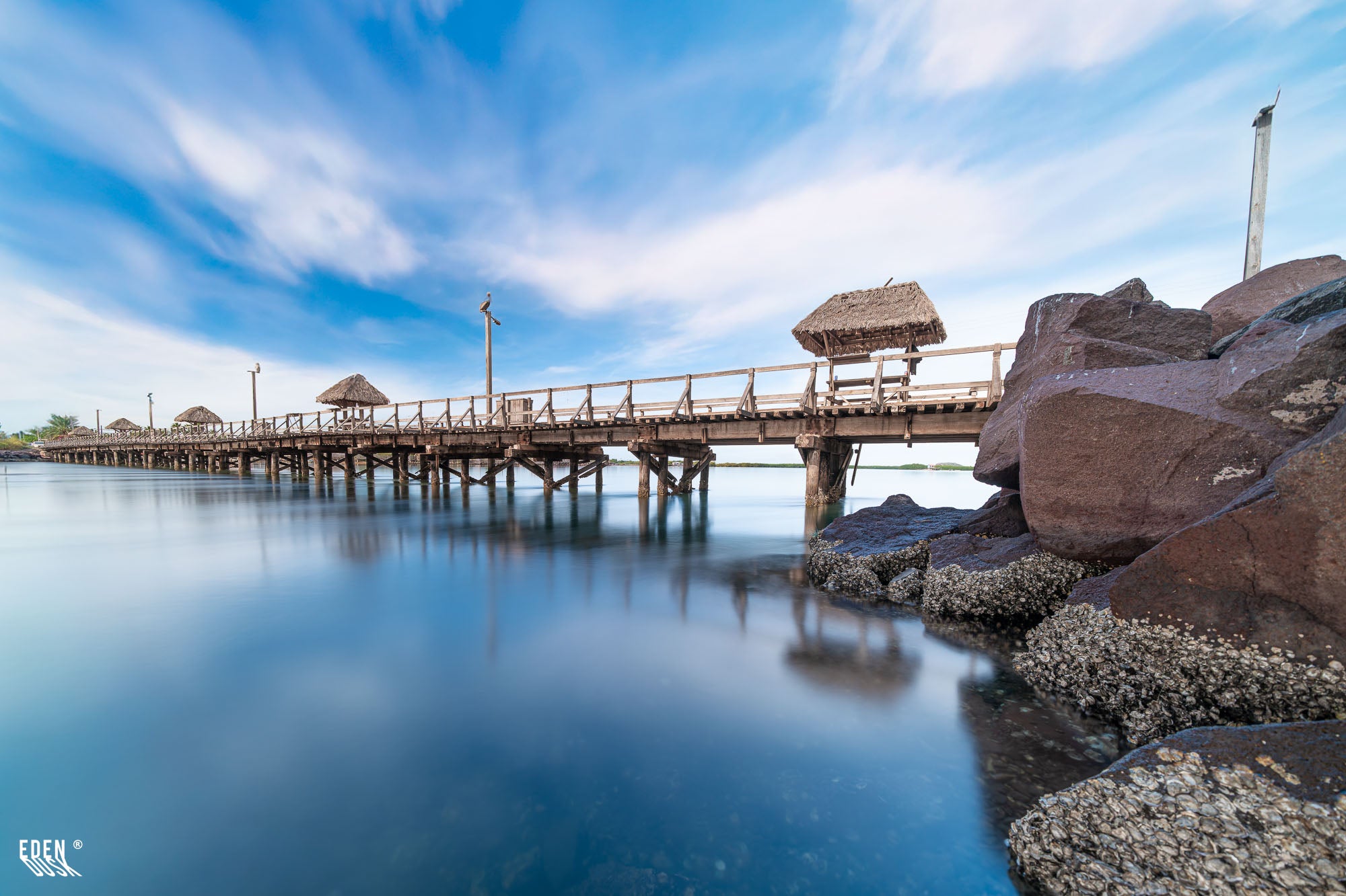 Puente de madera con palapas sobre agua tranquila; rocas con conchas en primer plano y cielo con nubes suaves.