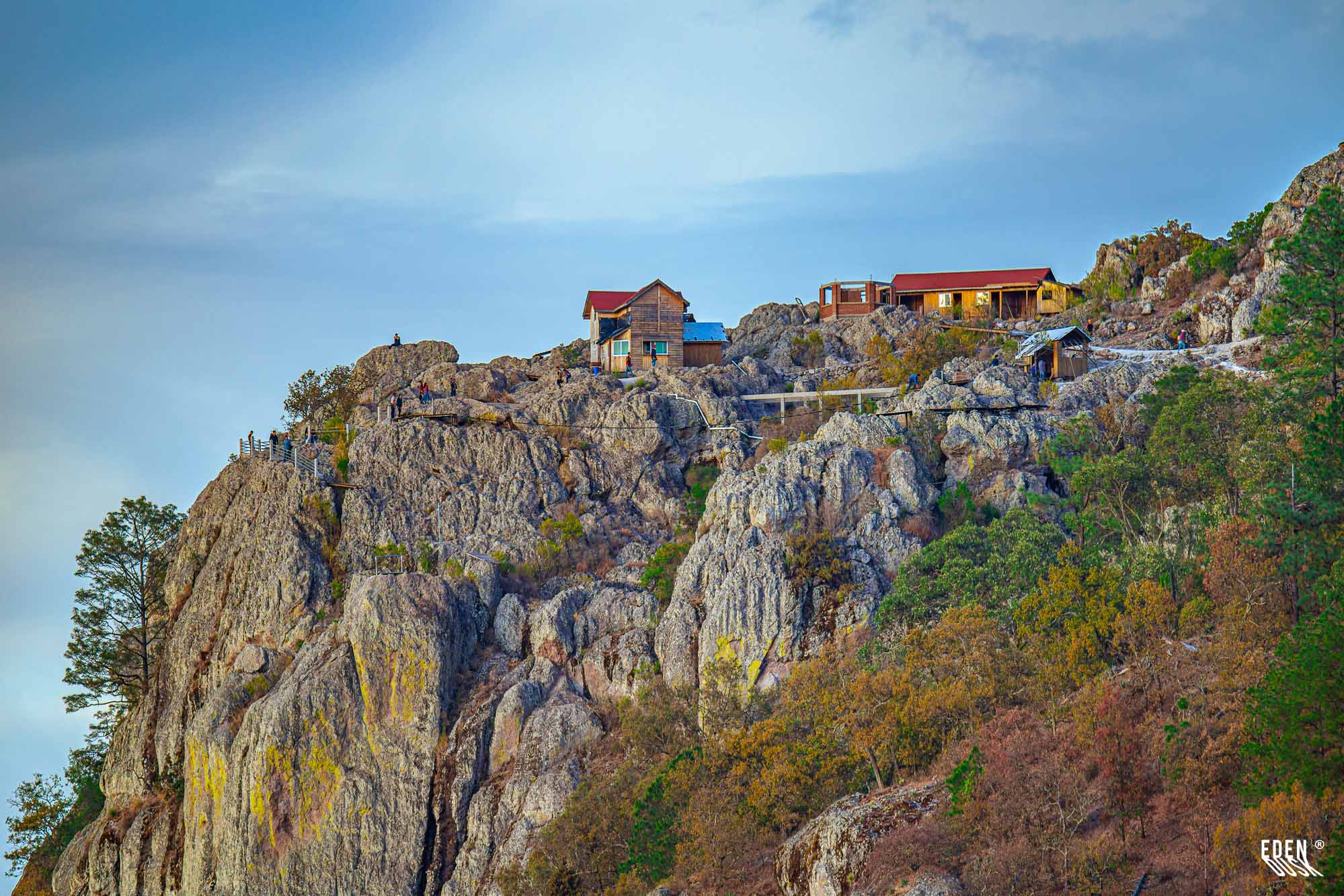 Cabañas de madera sobre un peñasco rocoso con barandales y pasarela; algunas personas en la cima y cielo azul claro.