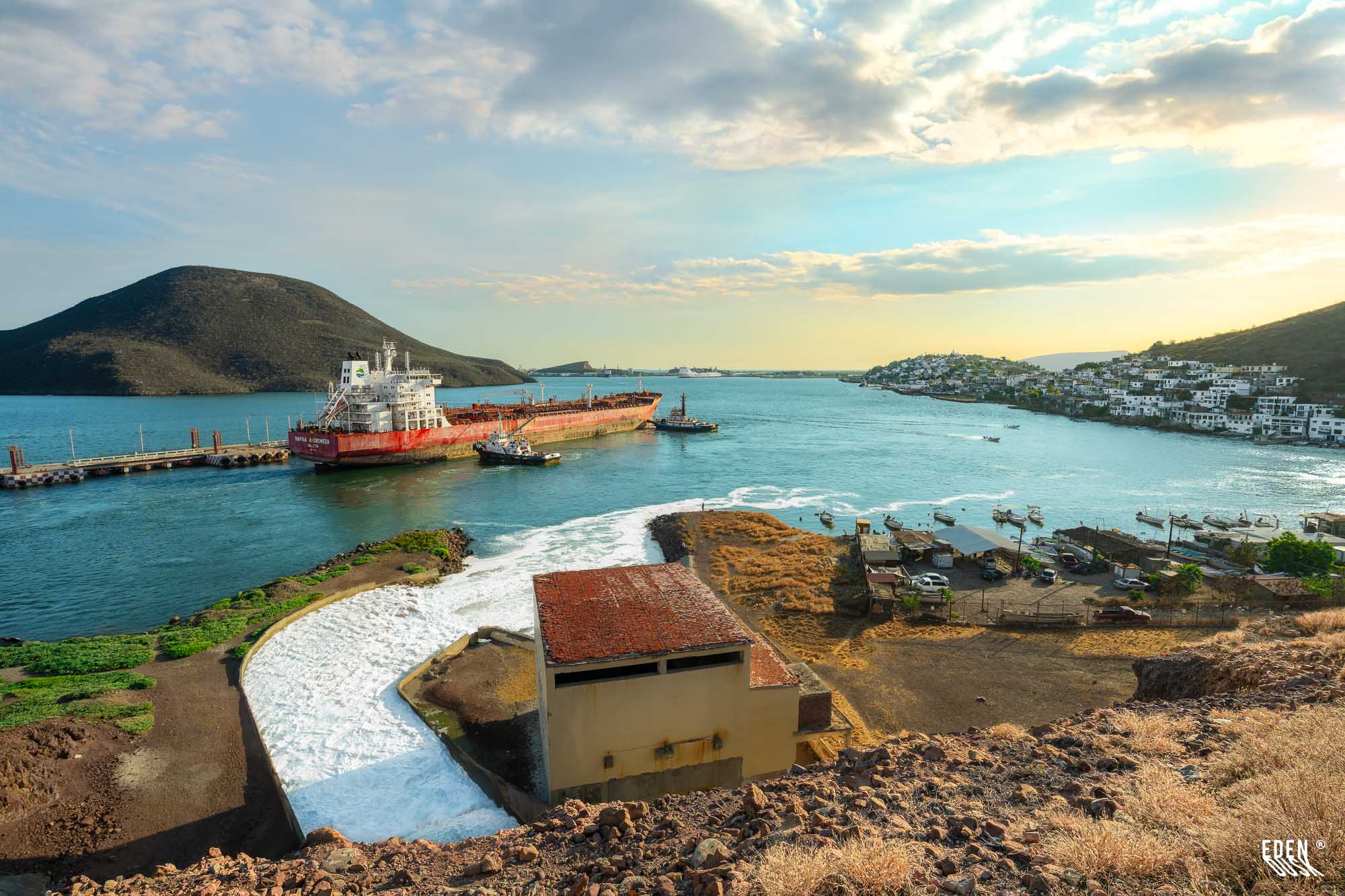 Vista panorámica de la bahía de Topolobampo al atardecer con buque rojo en el muelle, canal de espuma en primer plano y el pueblo blanco sobre la ladera.