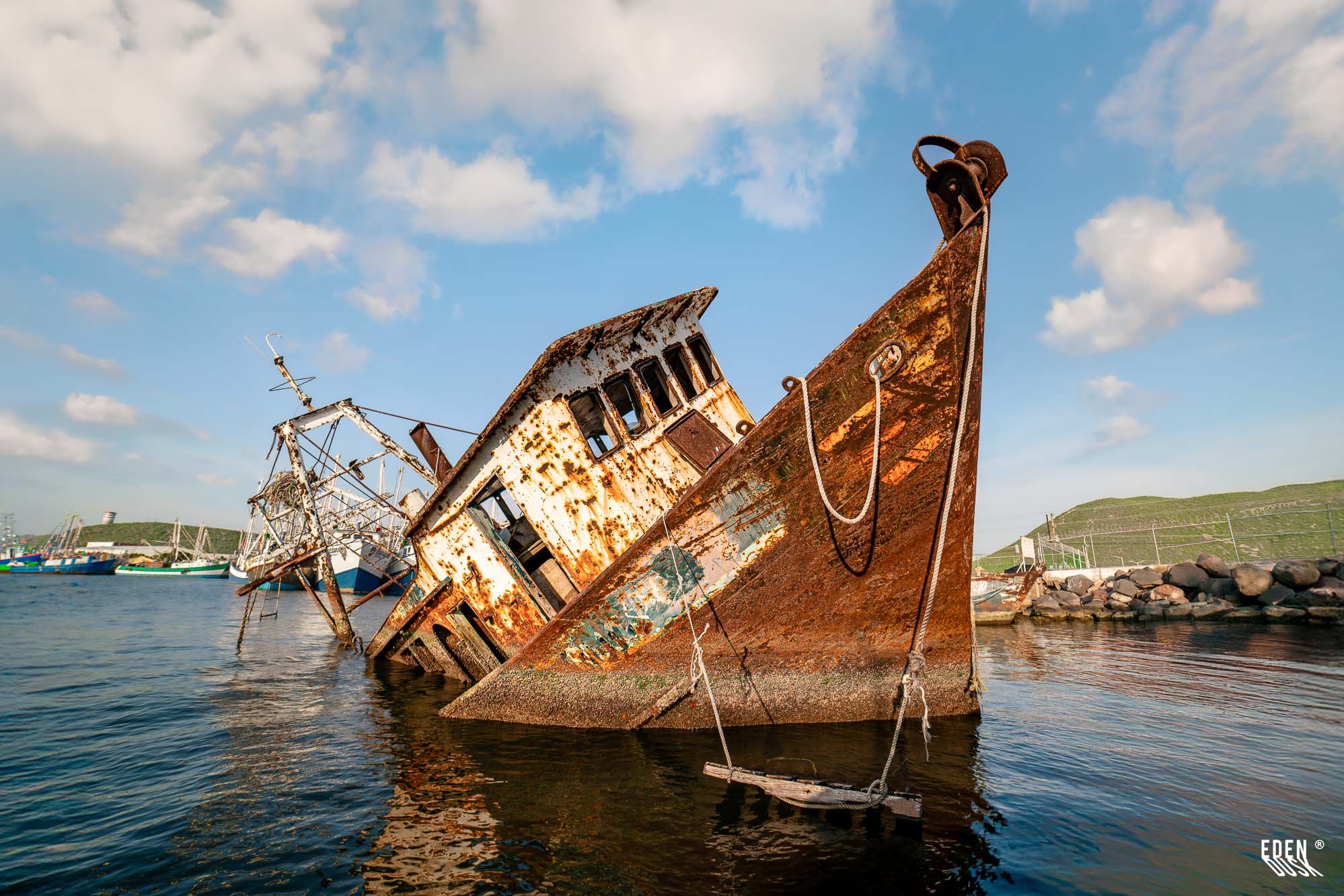 Barco camaronero oxidado y semihundido junto al muelle, con cielo azul y colinas verdes al fondo en el puerto de Topolobampo, Sinaloa.