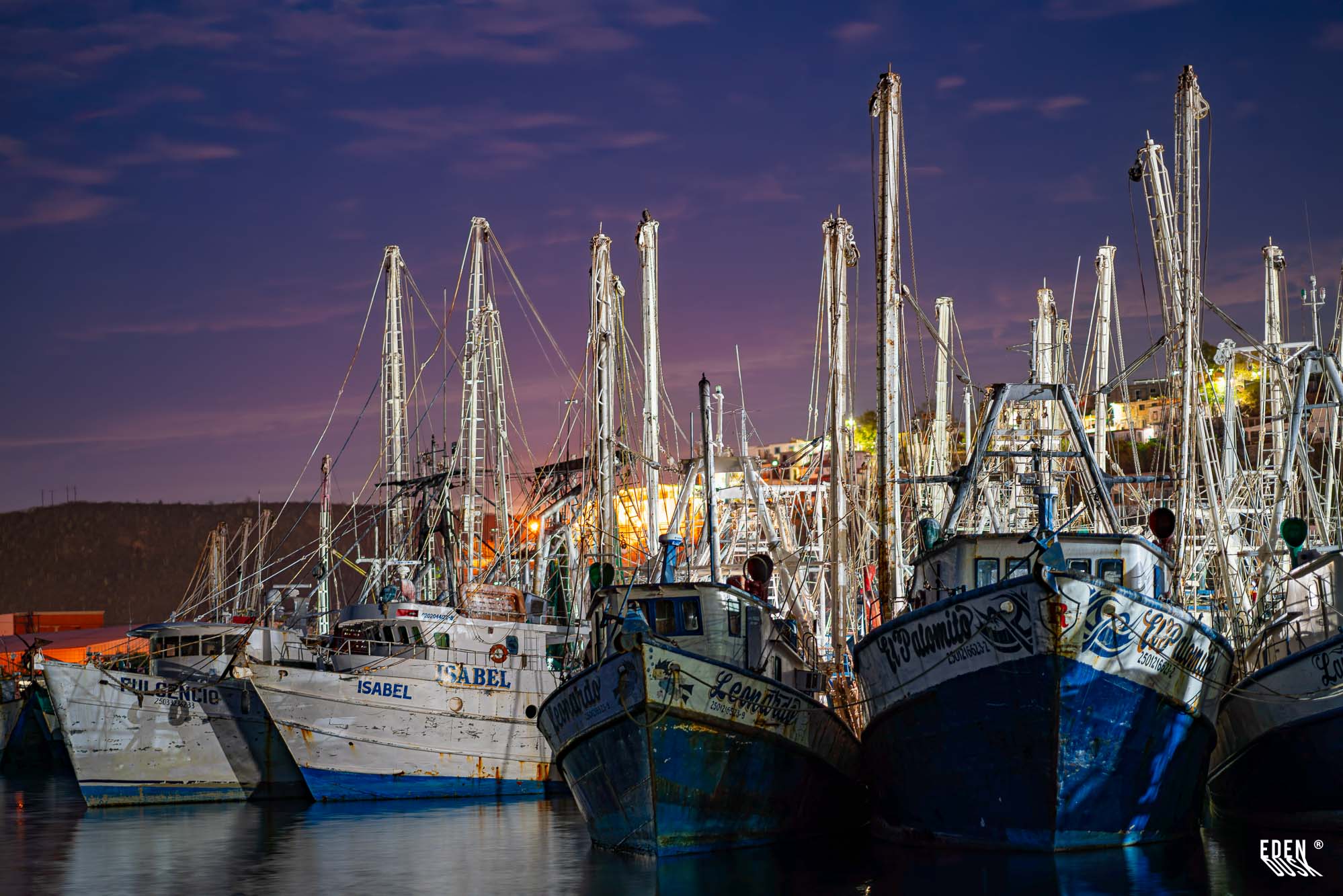 Barcos camaroneros alineados en el muelle al anochecer, mástiles y luces bajo cielo violeta, Topolobampo, Sinaloa.
