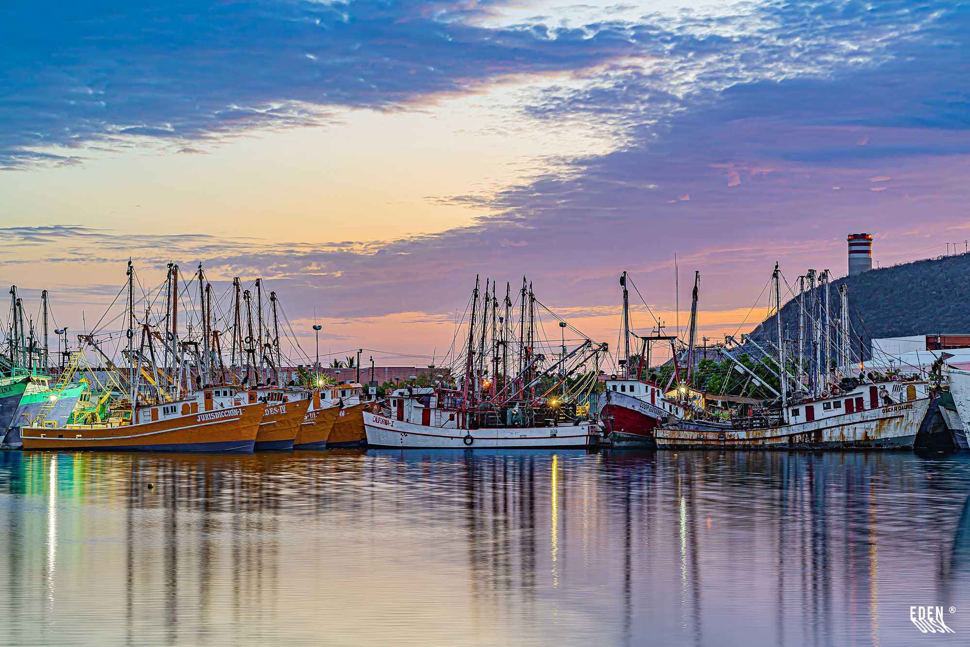 Flota camaronera alineada en el muelle al atardecer, cielo crepuscular y reflejos en el agua con torre en el cerro, Topolobampo, Sinaloa.
