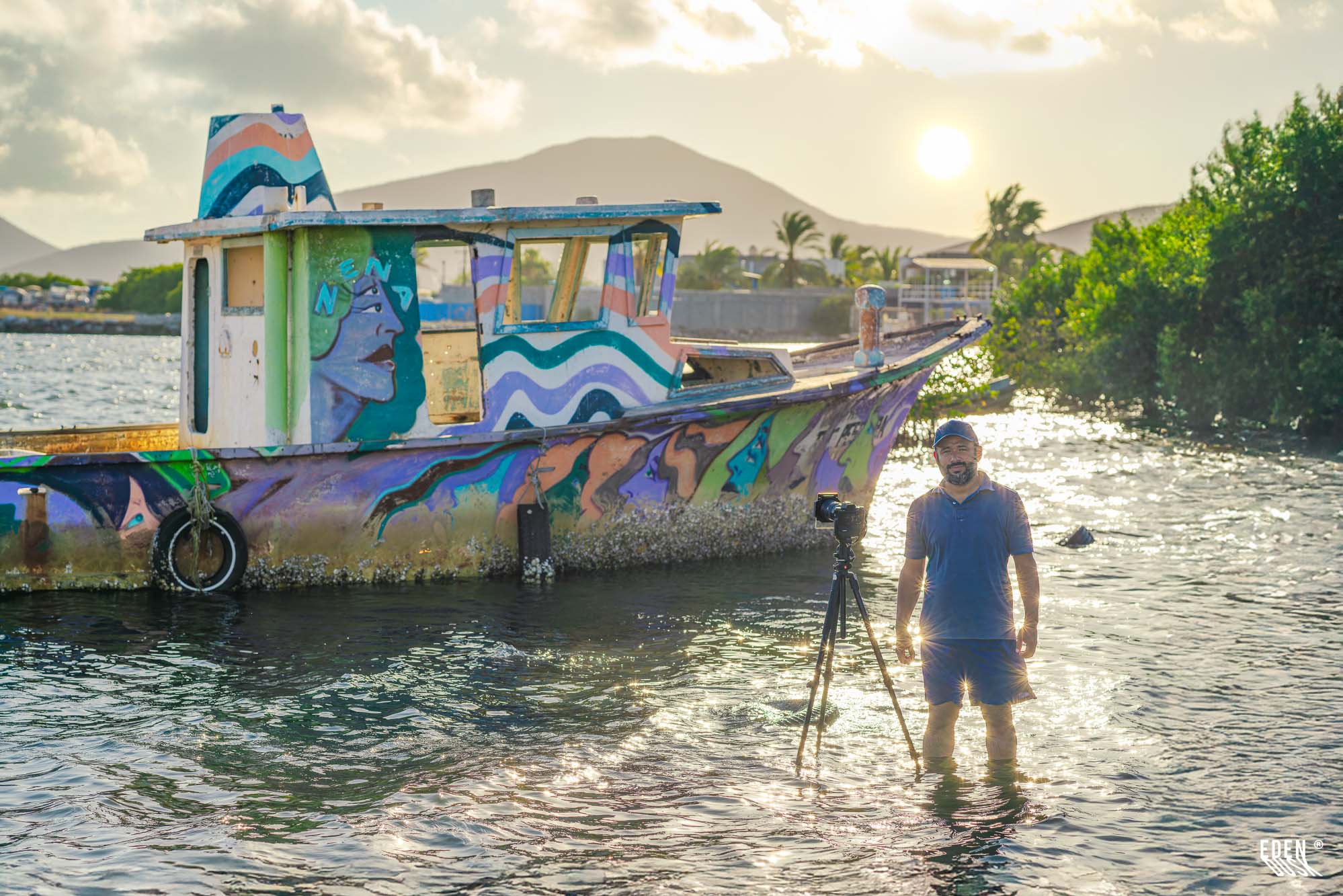 Fotógrafo de pie en el agua con trípode junto a lancha con grafiti, sol bajo y manglar al fondo, Topolobampo, Sinaloa.