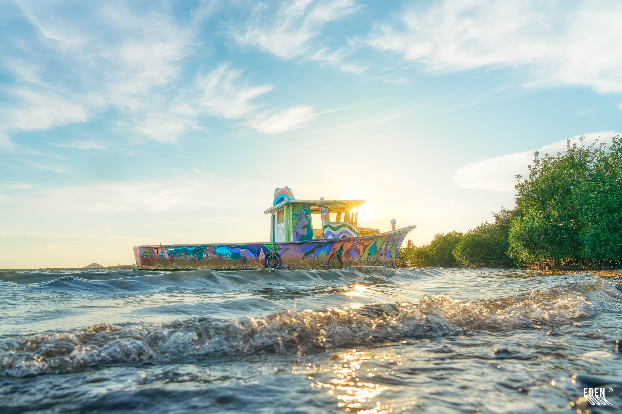 Lancha abandonada con arte grafiti junto a manglar, vista a ras del agua con pequeñas olas y sol poniente en Topolobampo, Sinaloa.