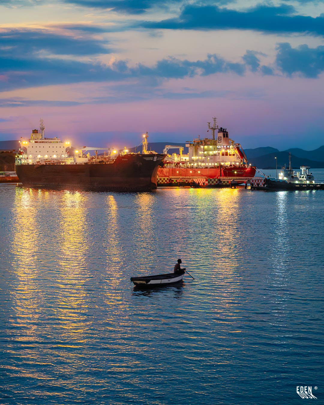 Pescador en lancha remando sobre aguas tranquilas con buques iluminados y reflejos al crepúsculo, Topolobampo, Sinaloa.