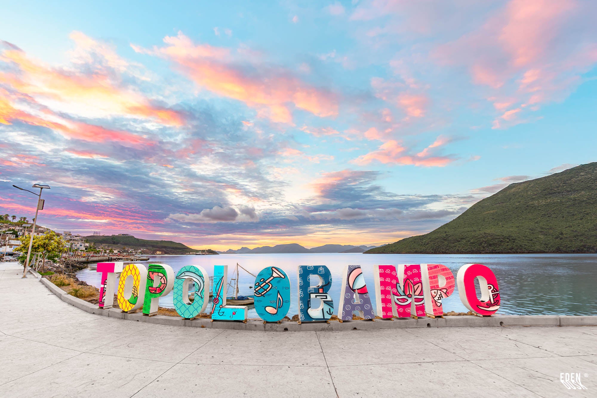 Letras coloridas de “Topolobampo” en el malecón, con bahía y cerros al fondo bajo nubes rosadas al amanecer, Sinaloa.