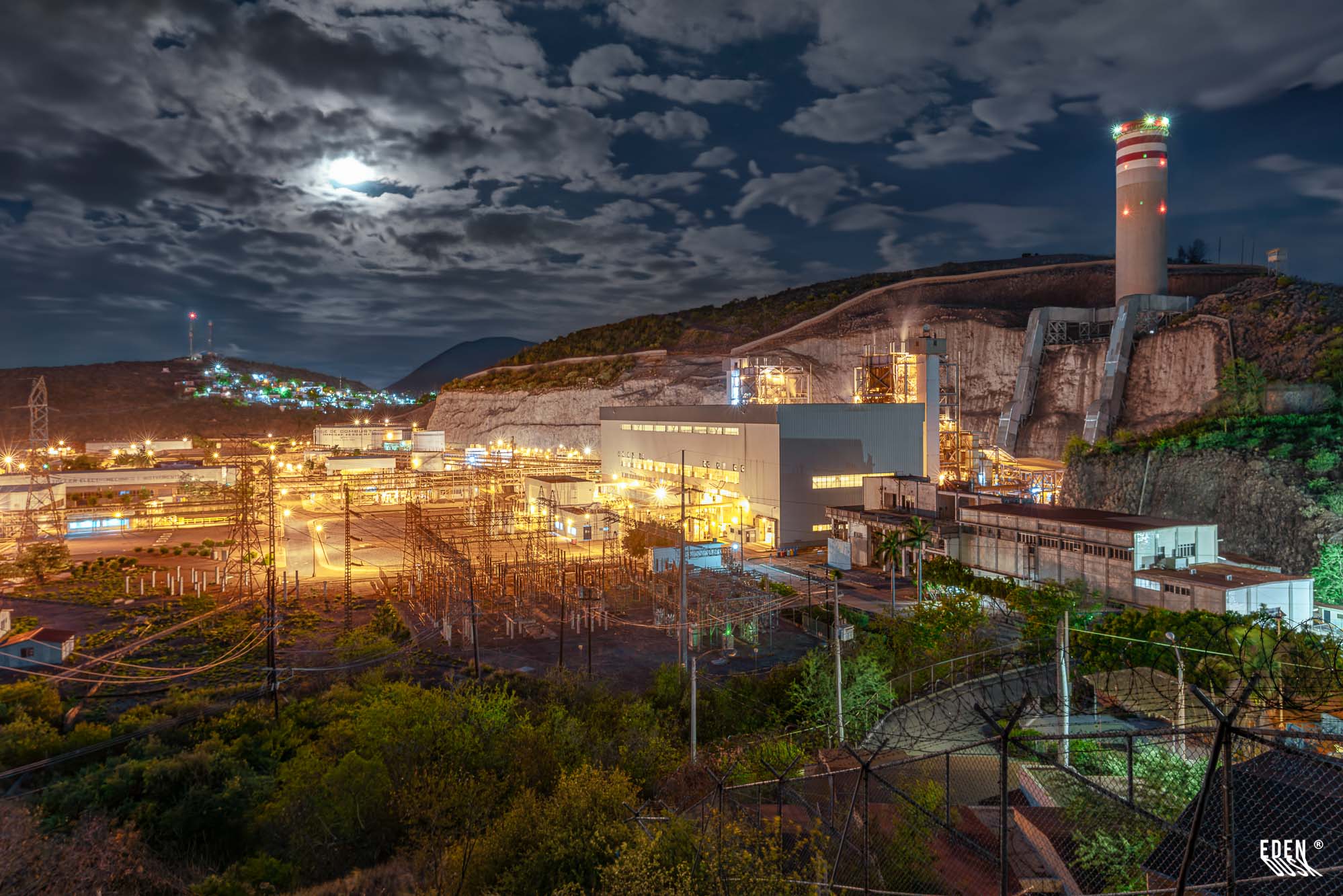 Planta industrial iluminada en Topolobampo con chimenea y subestación, luna entre nubes sobre las colinas y luces nocturnas que revelan la actividad del puerto, Sinaloa.